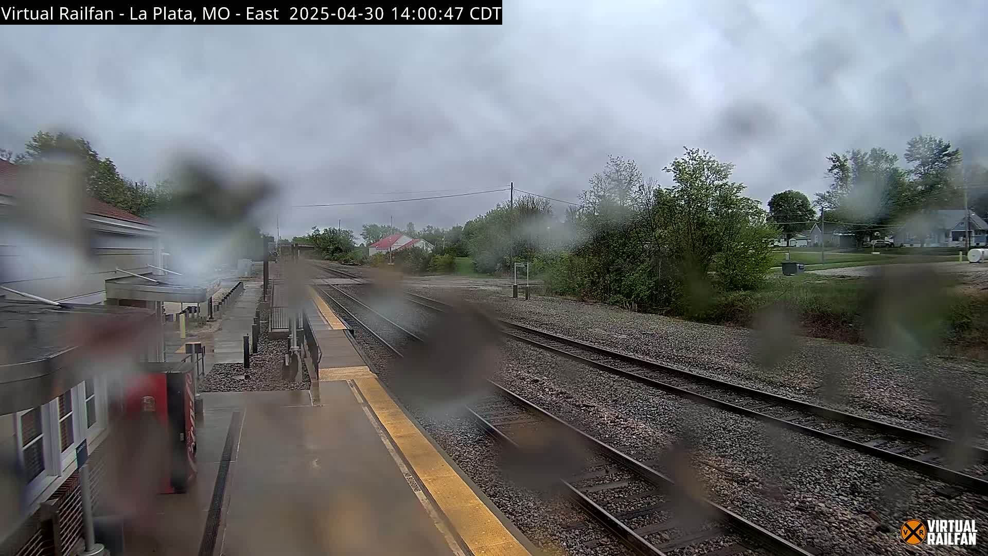 A rain-covered view of a train station platform and multiple train tracks, with trees and houses visible in the background under a cloudy sky.