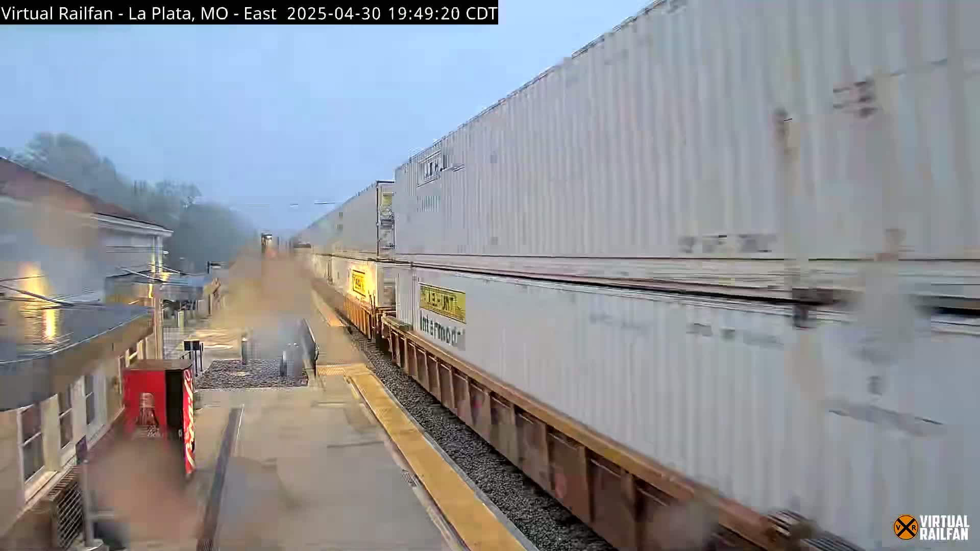 A long freight train carrying numerous white shipping containers passes a station on a rainy day.
