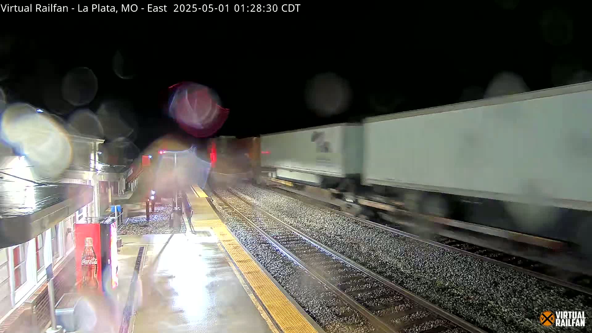 A long freight train speeds past a rain-slicked train platform at night.