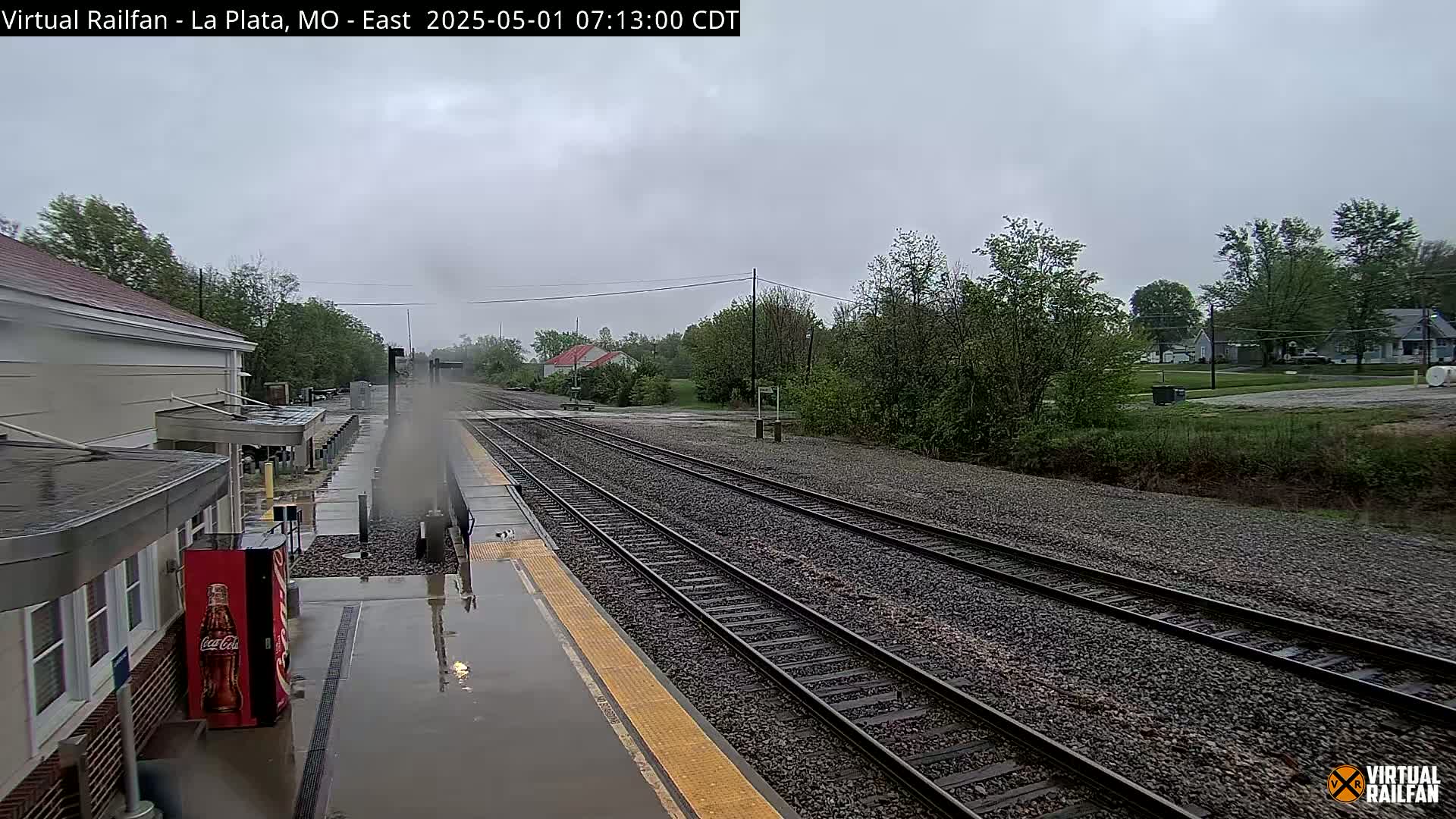 A rain-slicked train platform at a rural station, with multiple train tracks extending into the distance under an overcast sky.