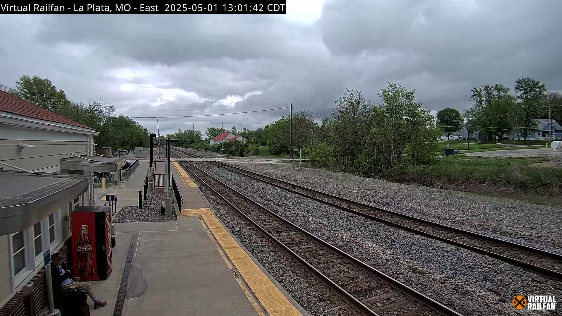 A mostly cloudy day at a train station shows multiple train tracks extending into the distance, with a person sitting near a Coca-Cola vending machine beside a building.