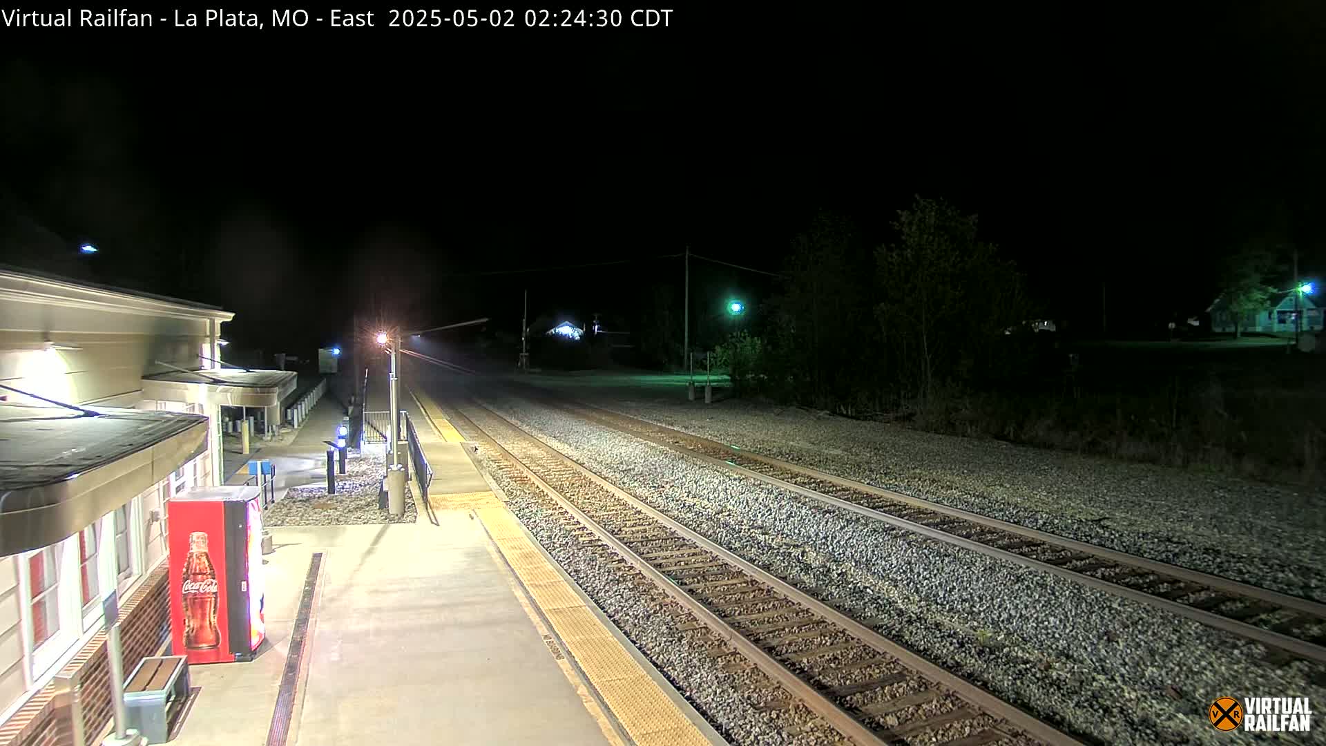A nighttime view of a train station platform with two sets of train tracks extending into the darkness, illuminated by artificial lights.