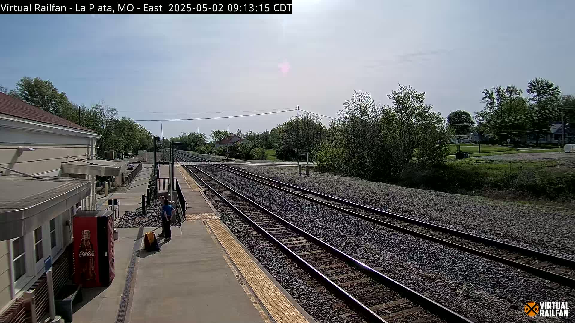 A train station platform with two people standing near a Coca-Cola vending machine, multiple train tracks extending into the distance under a mostly clear sky.