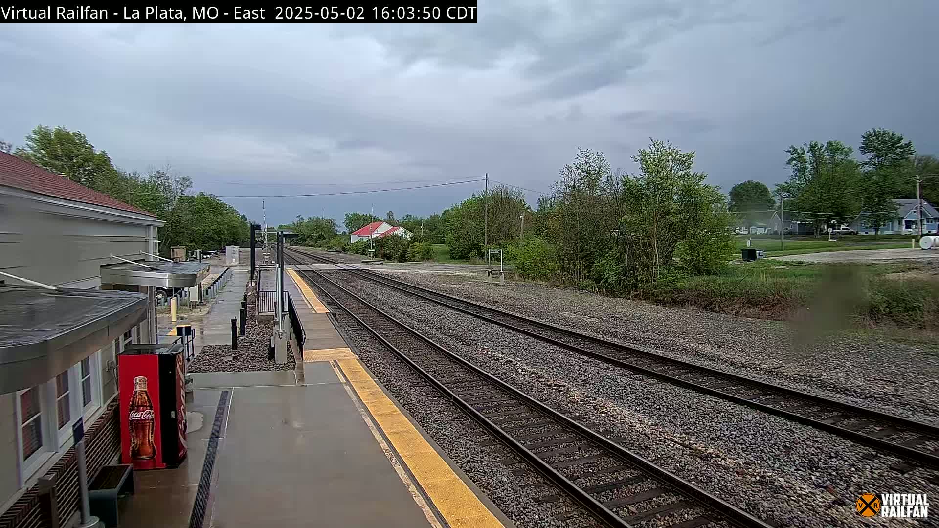 A wet train station platform overlooks several train tracks extending into a tree-lined landscape under a cloudy sky.