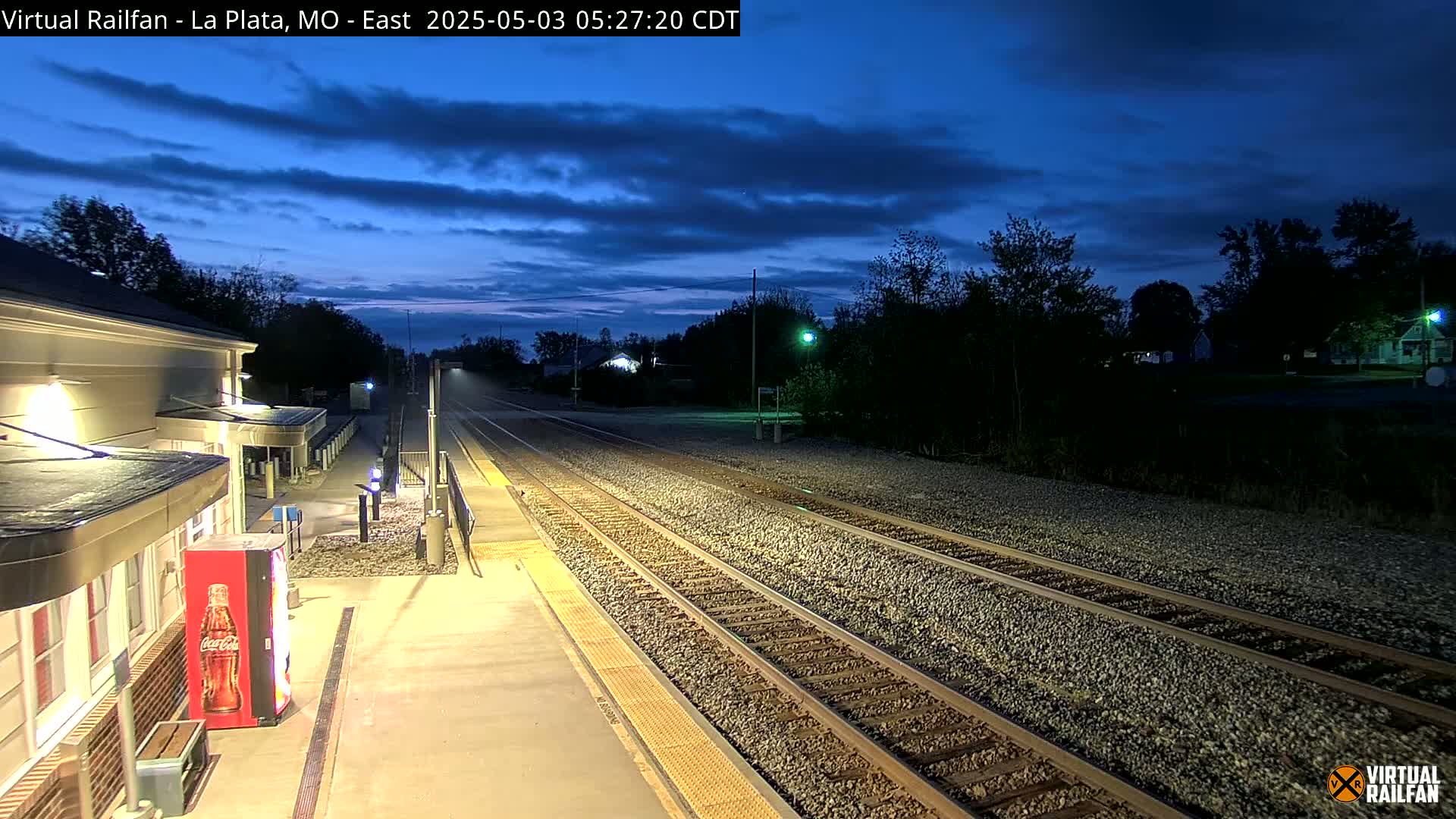 A train station platform at twilight, with train tracks extending into the distance under a mostly clear, dark blue sky.