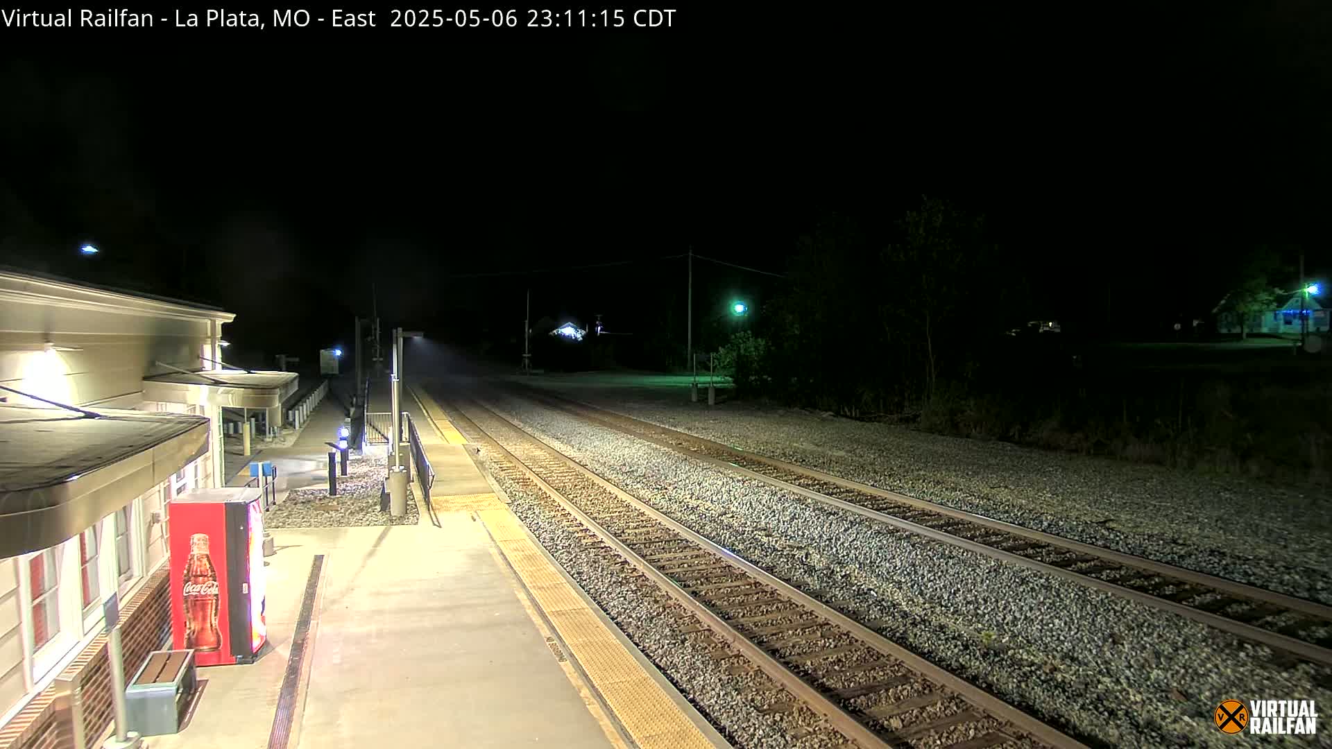 A nighttime view of a train station platform with a Coca-Cola vending machine, next to train tracks extending into the dark distance.