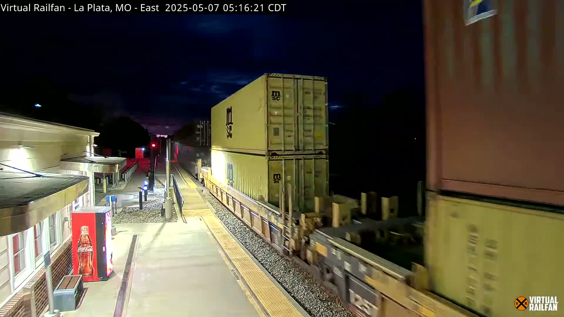 A freight train carrying stacked shipping containers passes a station at night under a mostly clear sky.