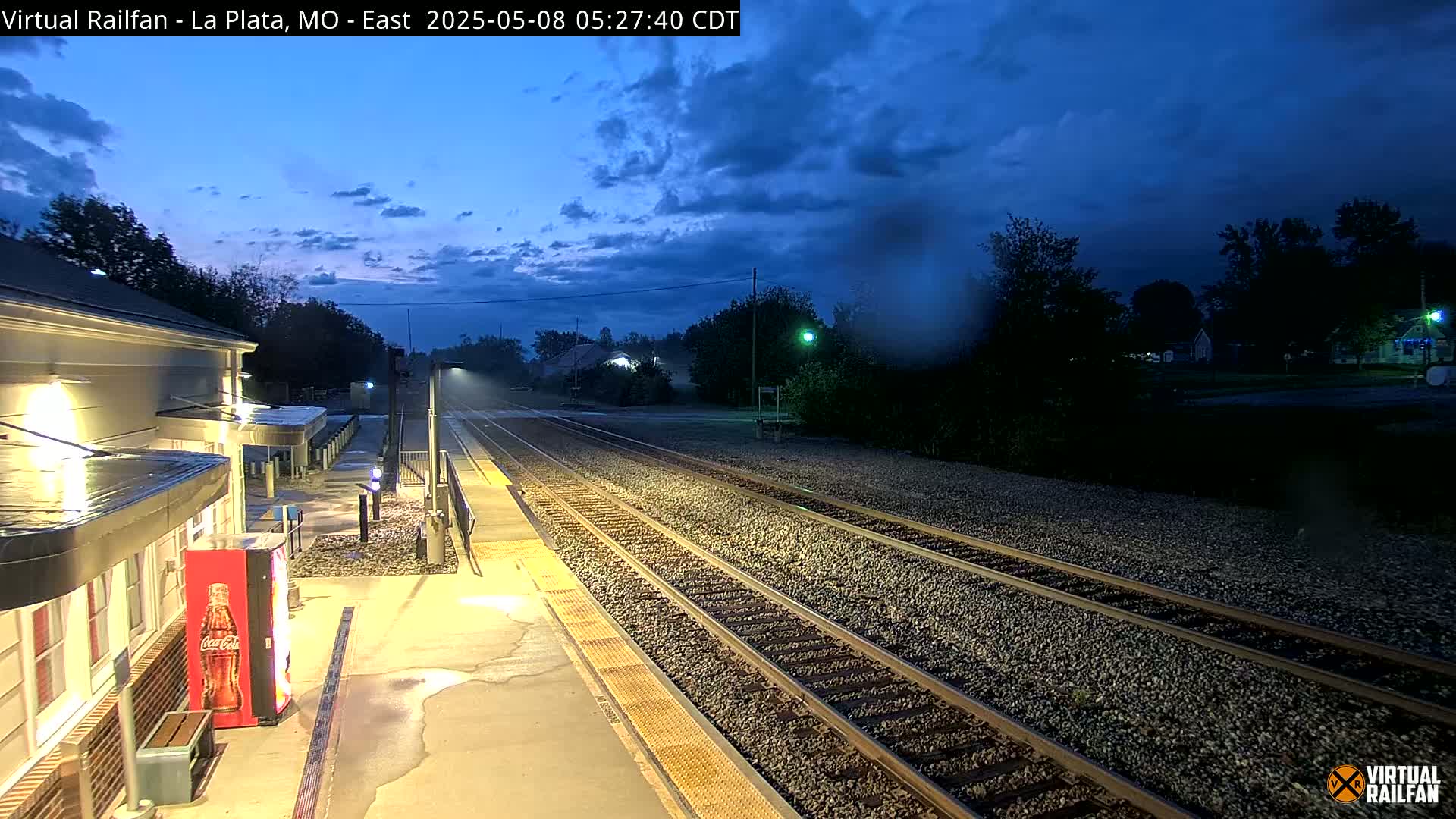 A train station platform at twilight, with multiple train tracks extending into the distance under a partly cloudy sky.