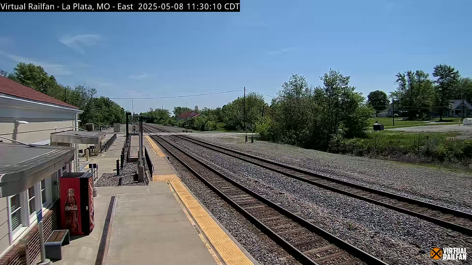 A train station platform on a sunny day, with multiple train tracks extending into the distance, surrounded by green trees and vegetation.
