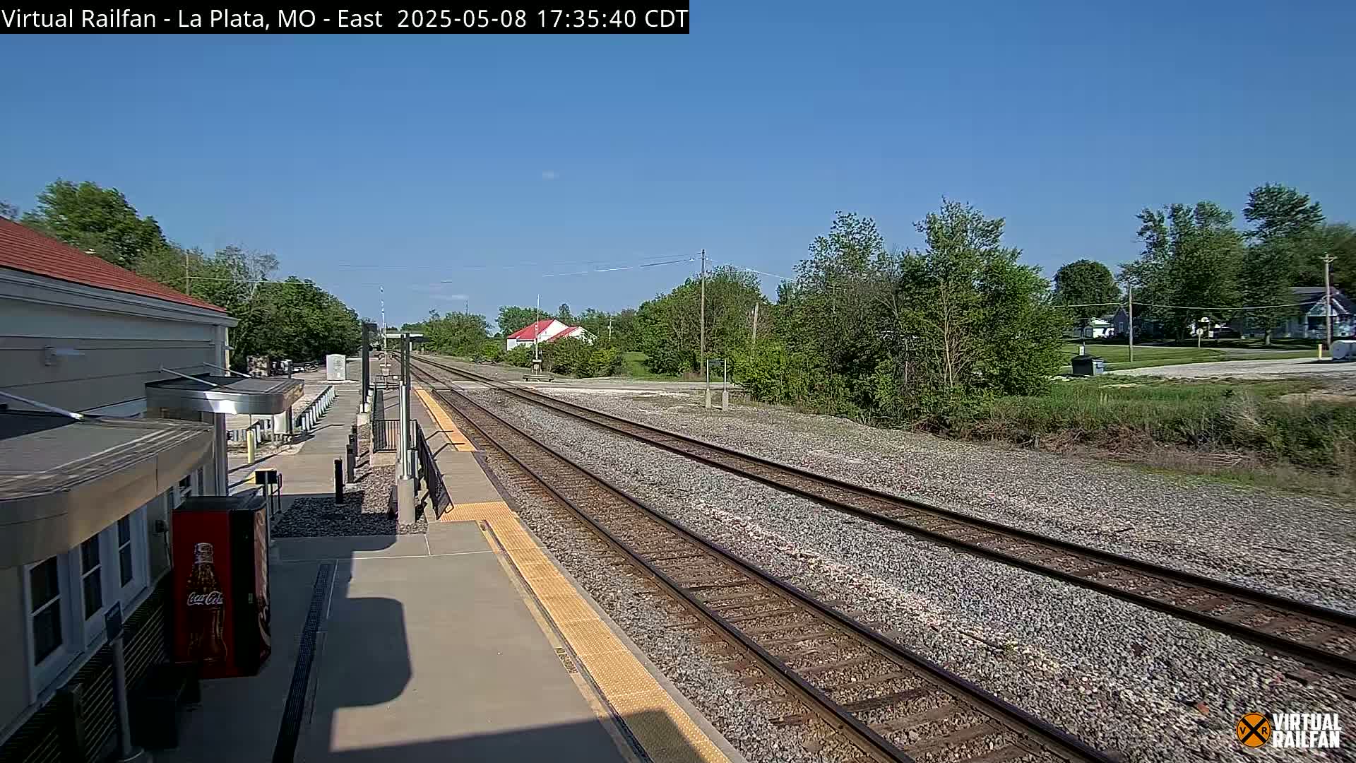 A train station platform with two sets of train tracks extending into a sunny, tree-lined landscape.