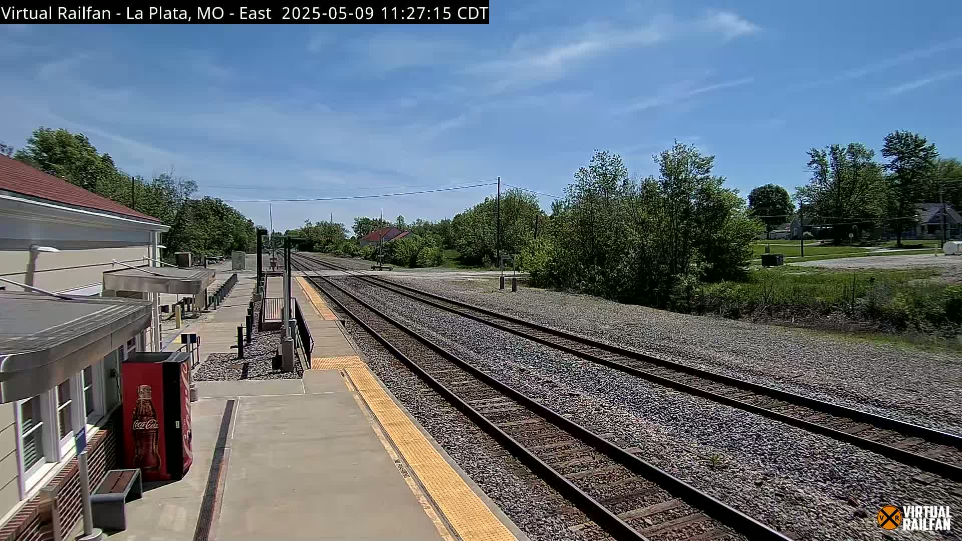A train station platform with multiple train tracks stretches into the distance under a mostly sunny sky.