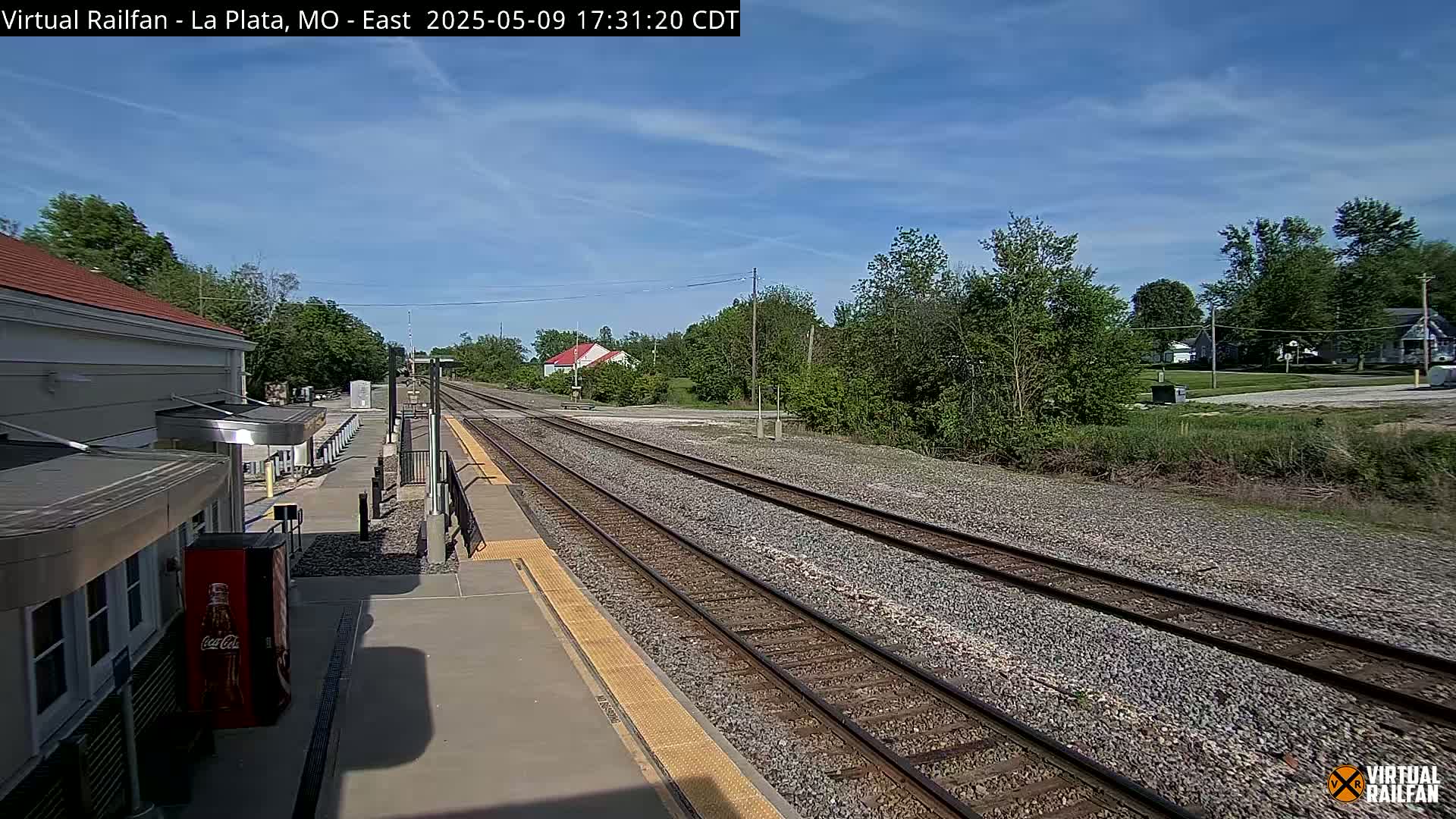 A train station platform with multiple train tracks extending into the distance under a mostly sunny sky.