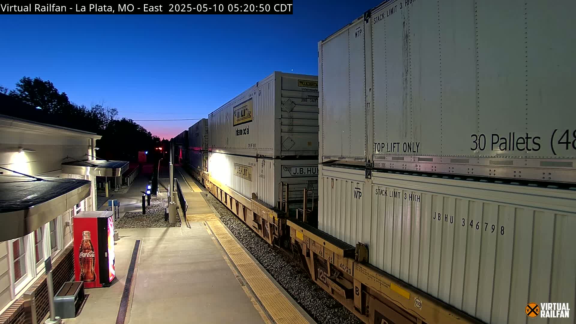A freight train carrying multiple shipping containers is passing through a train station at twilight.