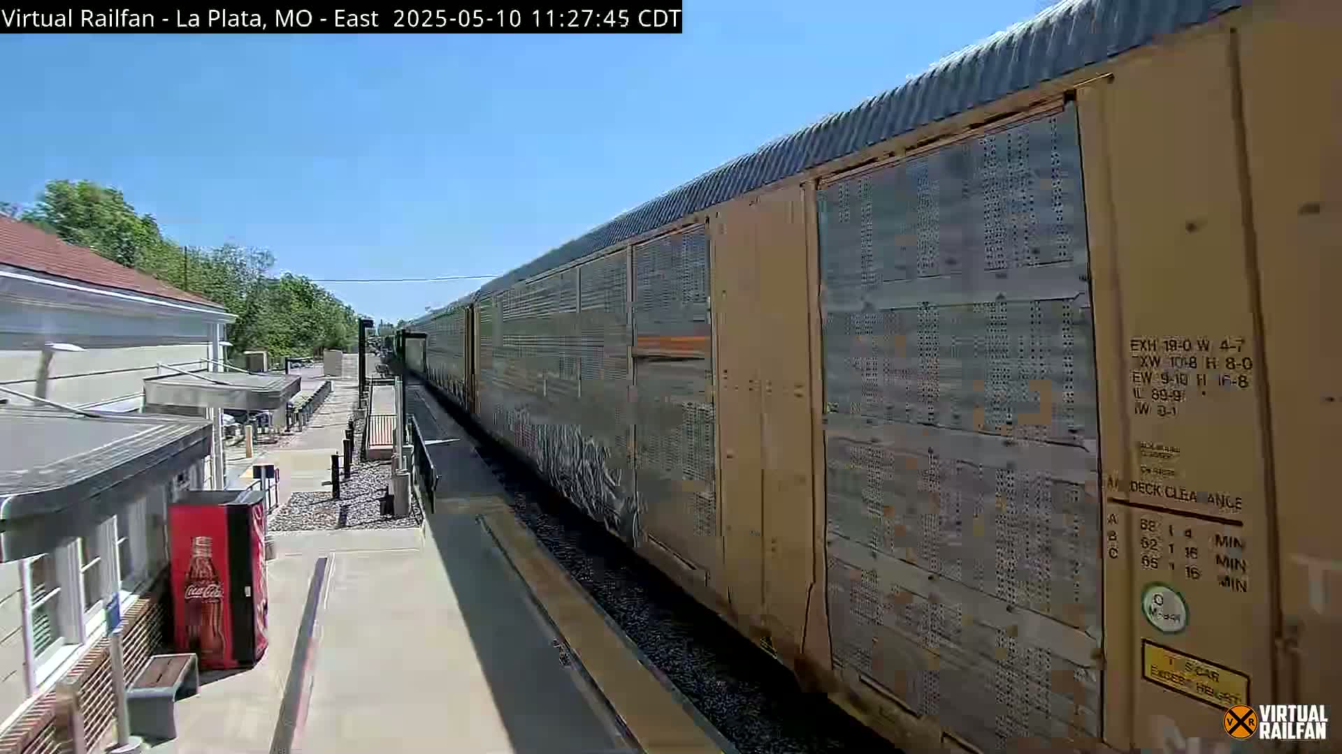 A long freight train passes a small train station on a sunny day.