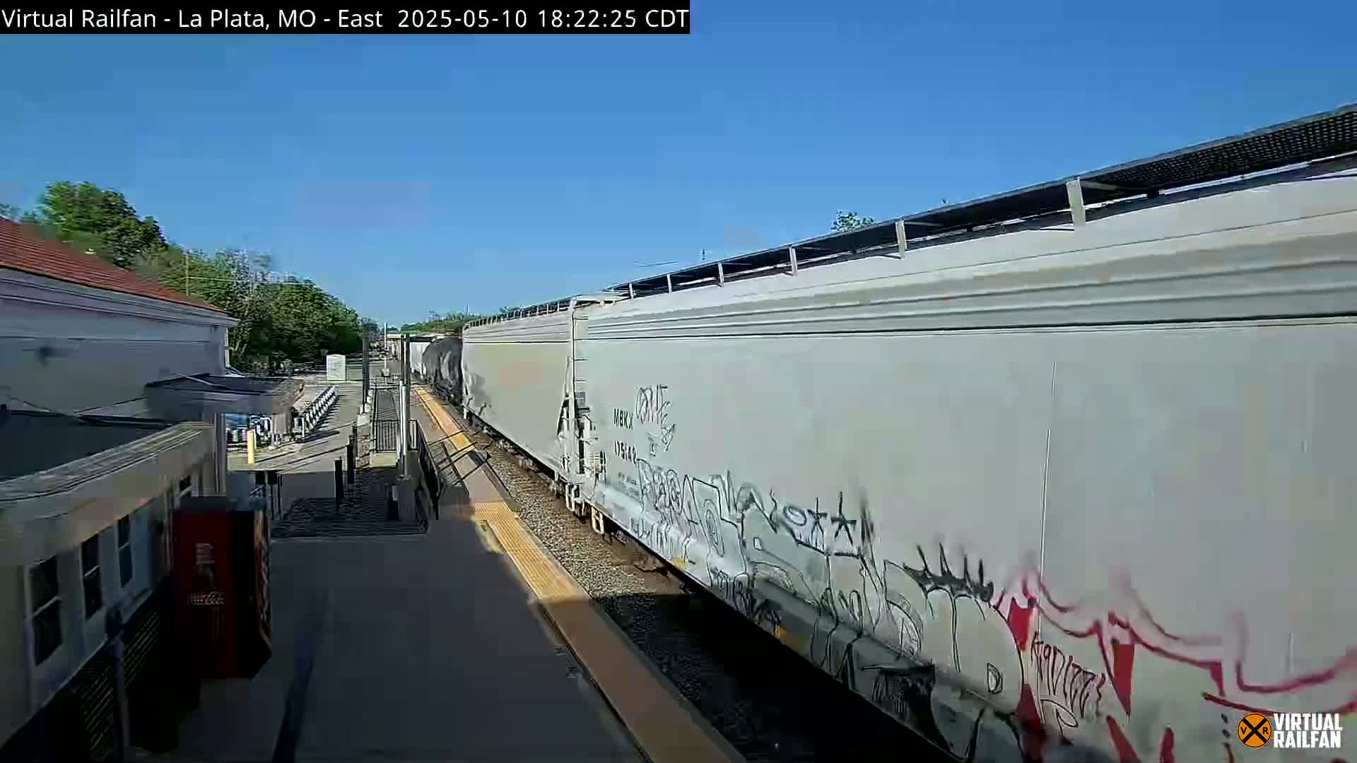 A long freight train with graffiti on its cars passes a train station on a sunny day.