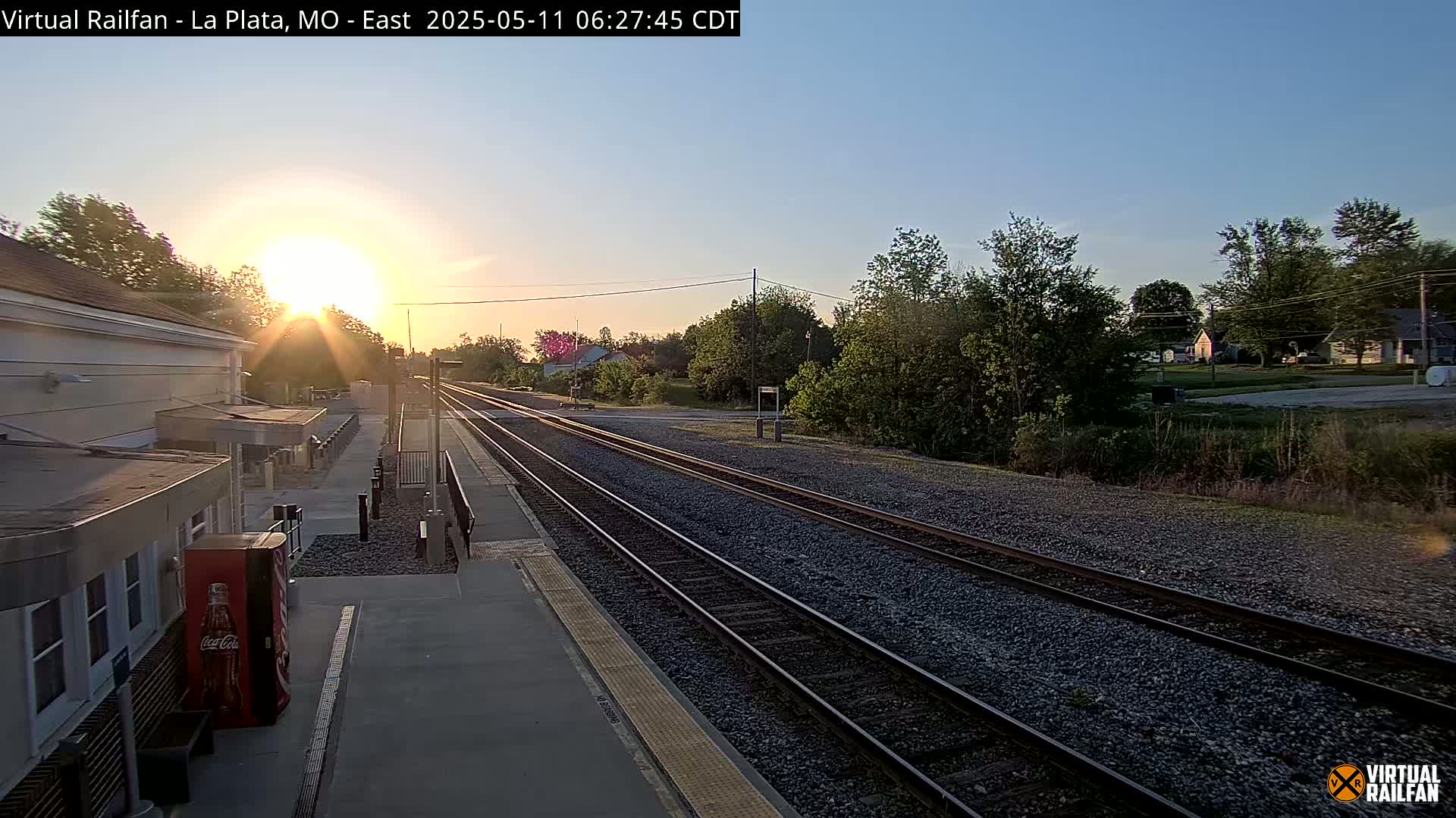 A sunrise illuminates a train station platform and tracks extending into a small town under a clear sky.