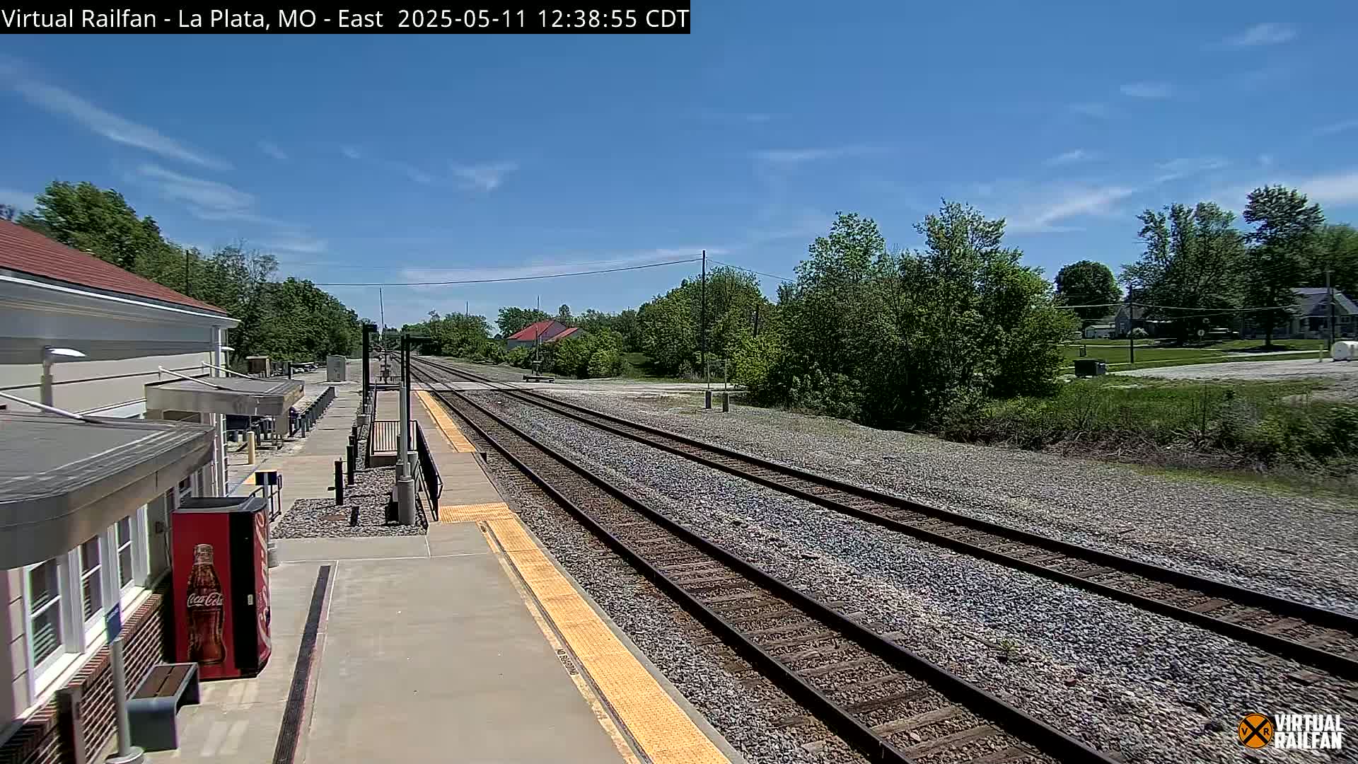 A train station platform with a building and multiple train tracks extends into a sunny, tree-lined landscape under a mostly clear blue sky.