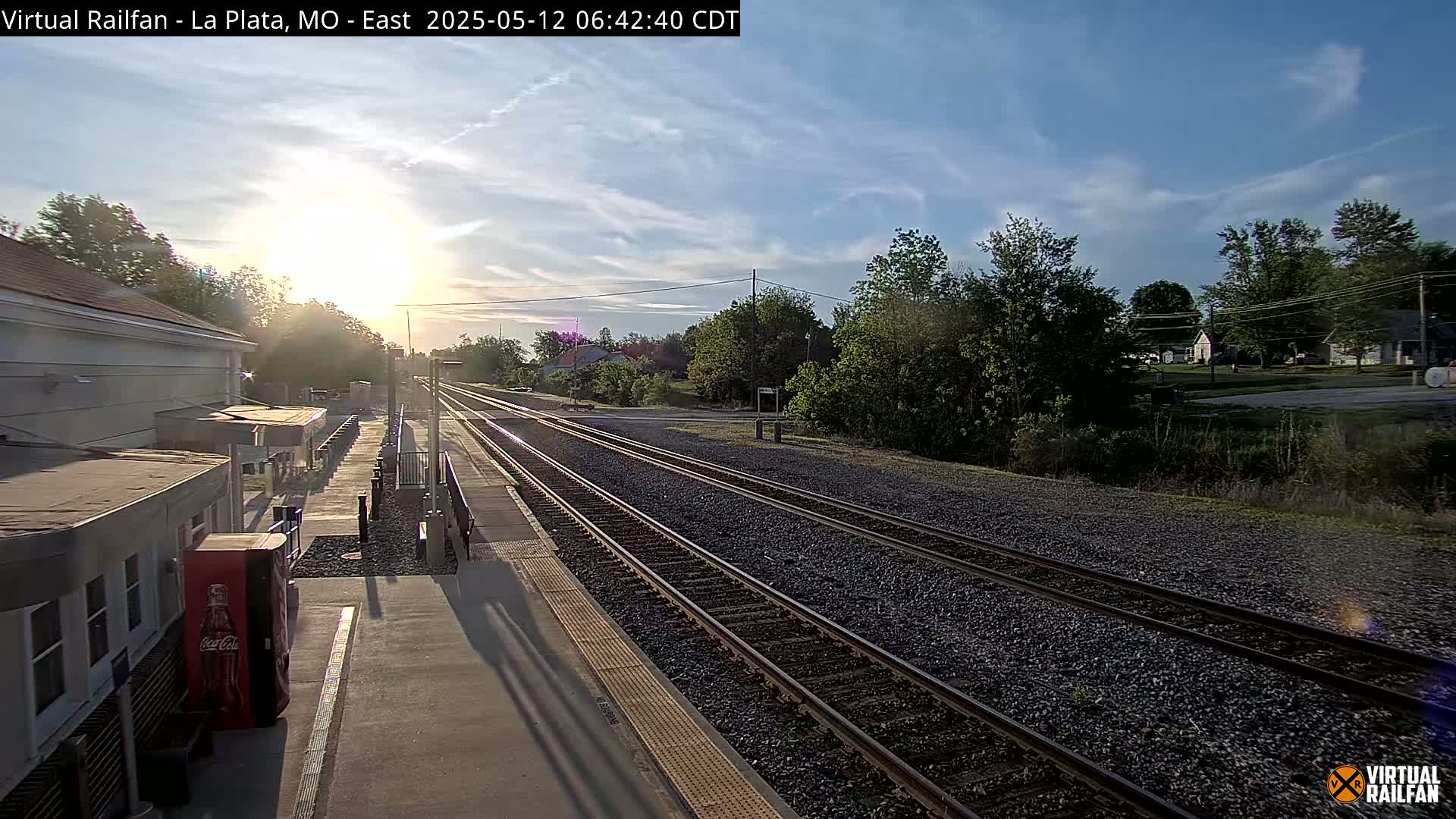 A train station platform with multiple tracks extends into a sunlit landscape of trees and houses under a mostly clear sky.
