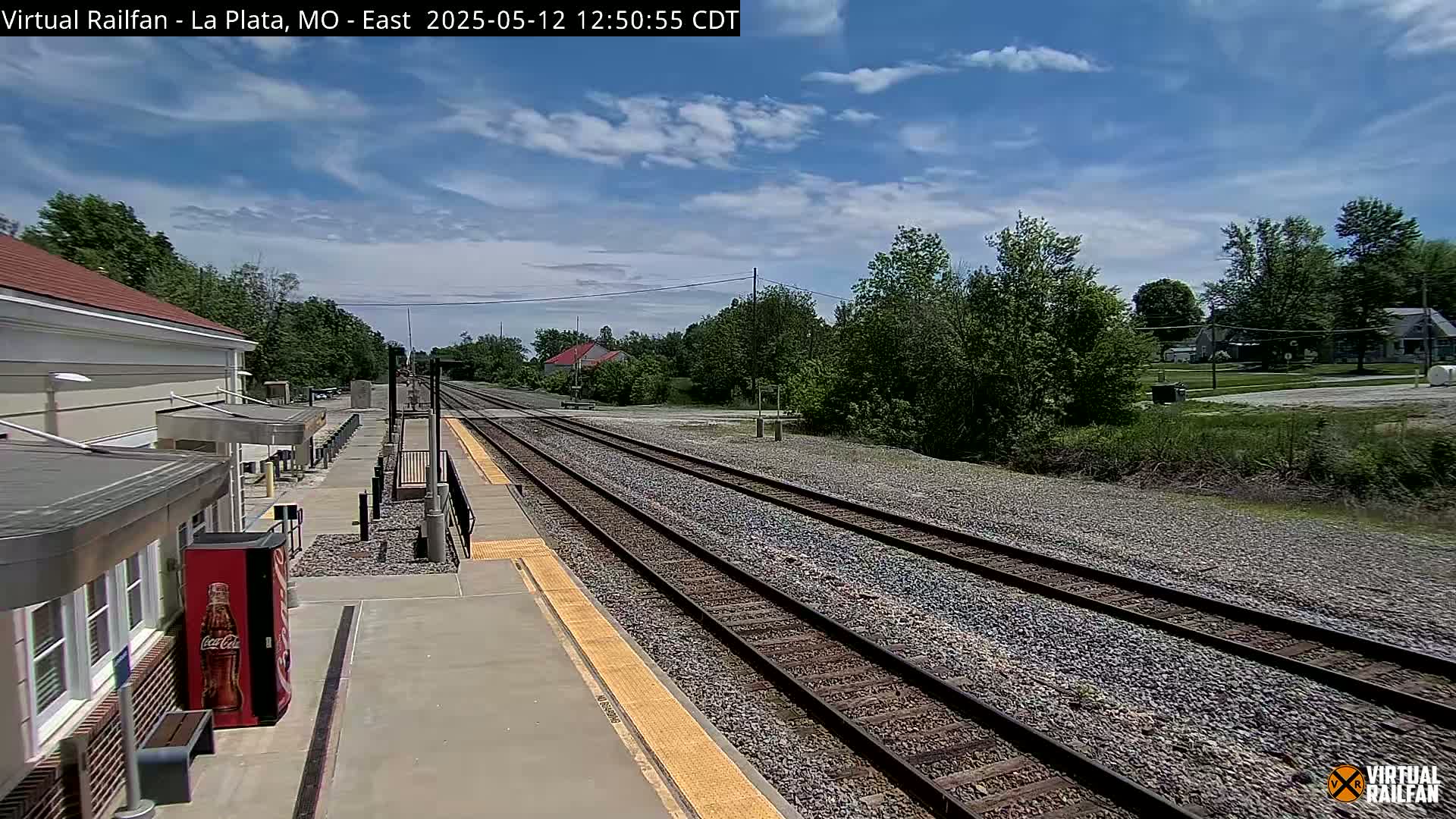 A mostly sunny day at a train station with multiple tracks extending into the distance, surrounded by trees and a few buildings.