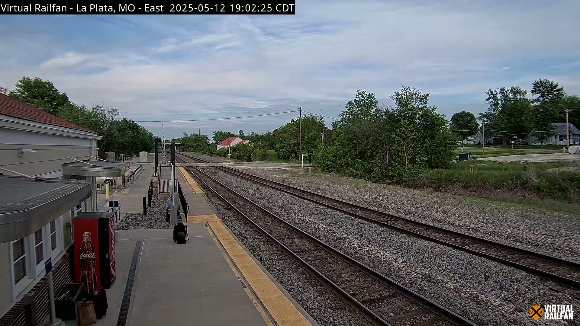 A mostly cloudy daytime view of a train station platform and multiple train tracks extending into the distance, surrounded by green trees and houses.