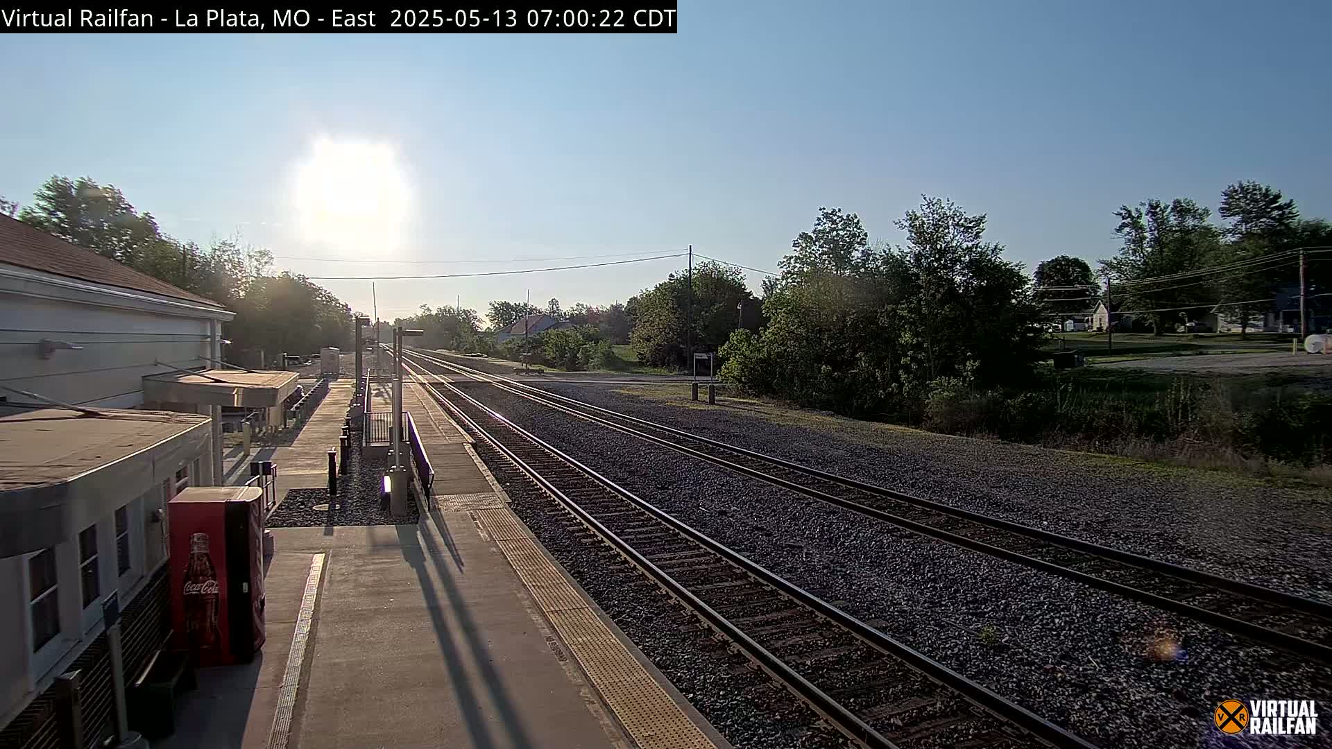 A sunny morning view of a train station platform with multiple train tracks extending into the distance, surrounded by trees and buildings.