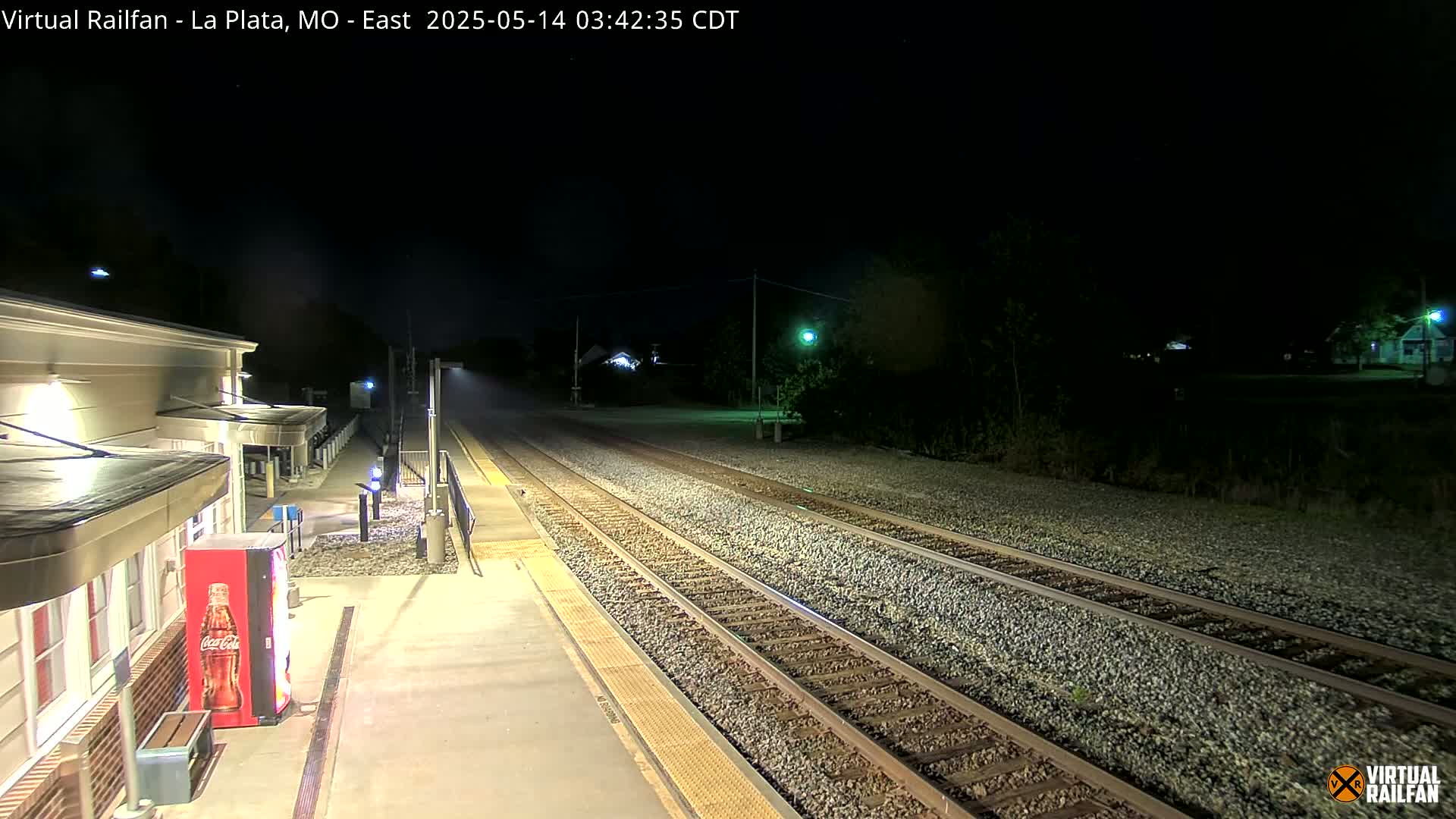 A nighttime view of a train station platform and railway tracks, illuminated by artificial lights.