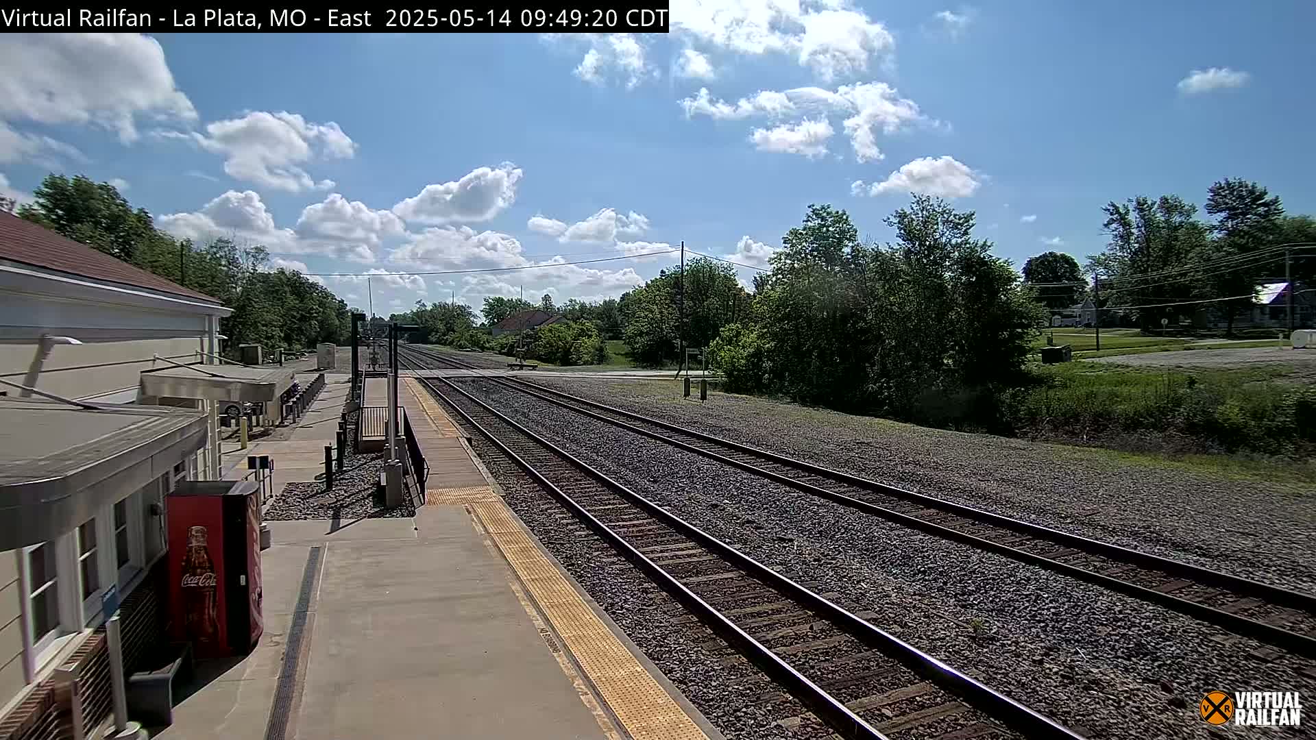 A mostly sunny day at a train station with multiple train tracks extending into the distance, surrounded by trees and a few buildings.