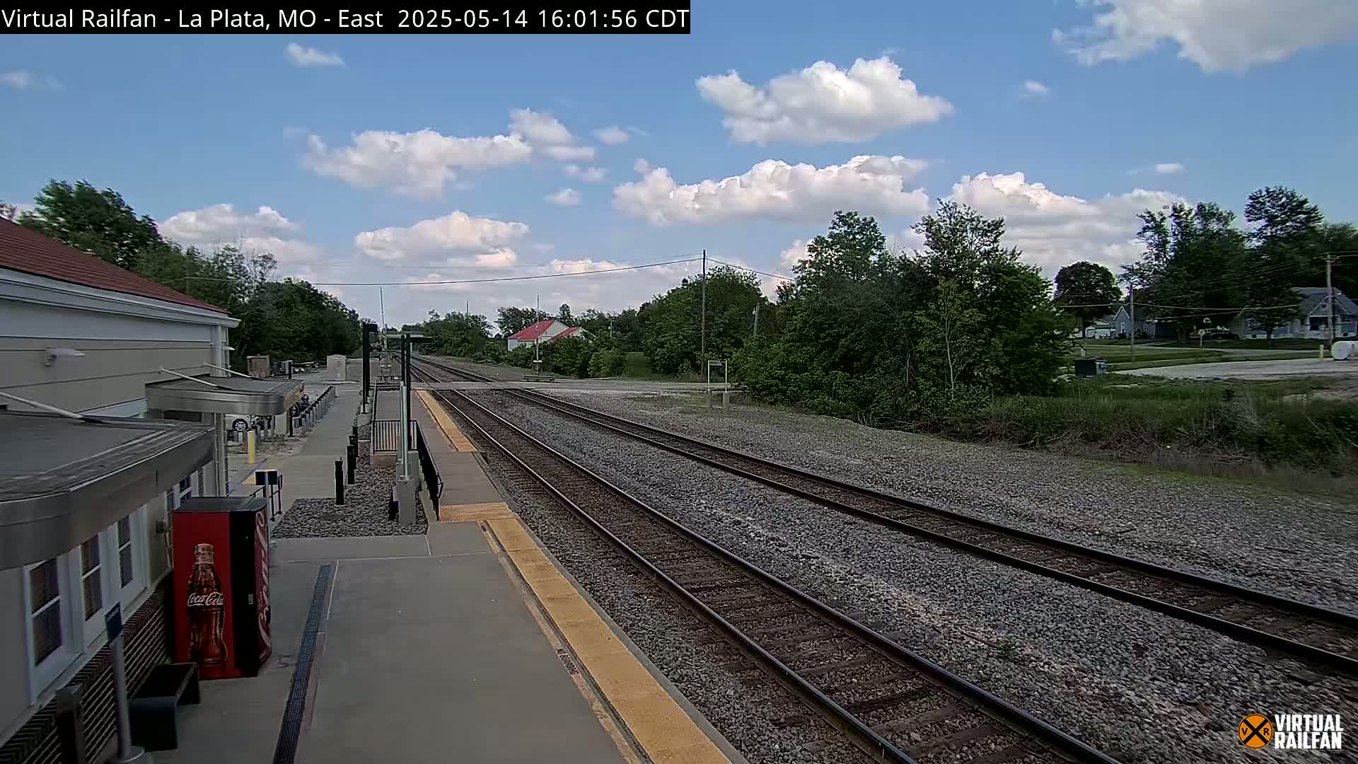 A train station platform with multiple tracks extends into a partly cloudy landscape, featuring a building and surrounding vegetation.
