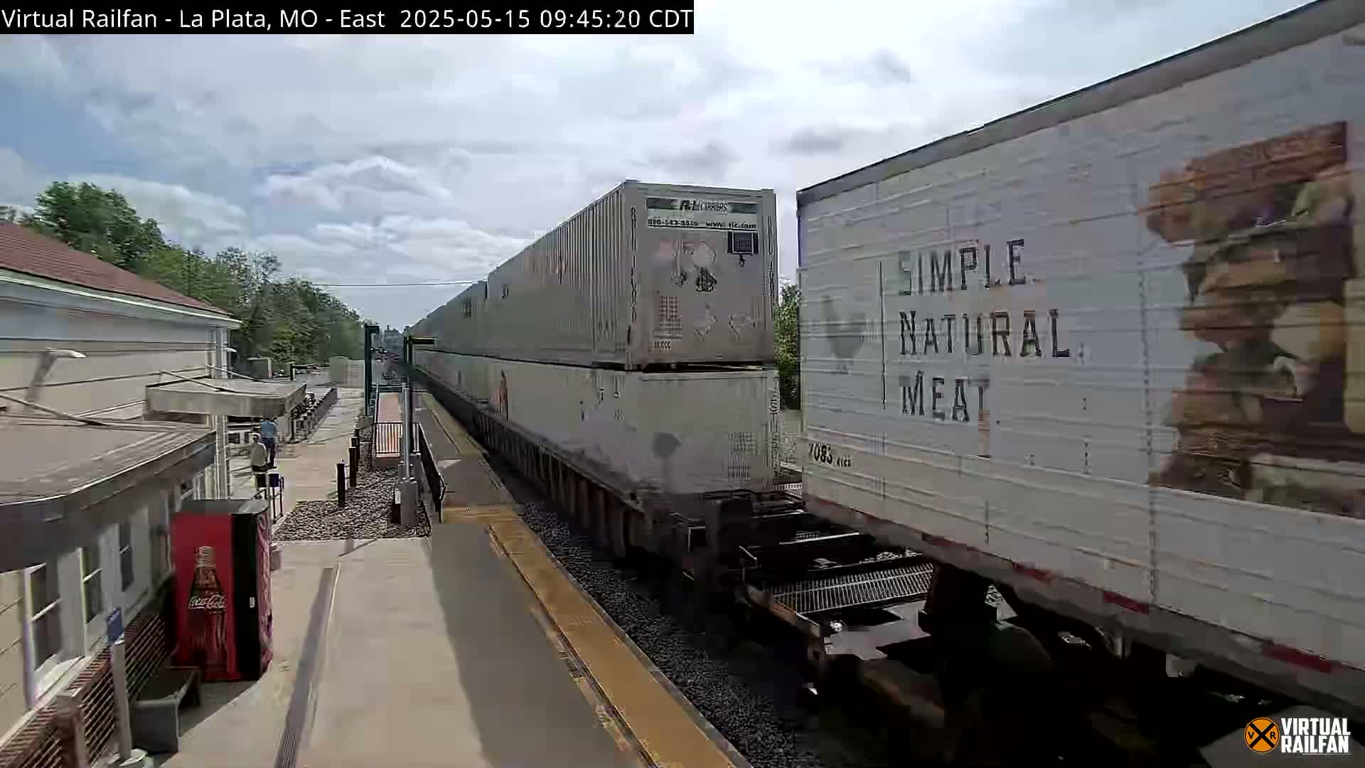 A long freight train carrying stacked containers passes a train station on a partly cloudy day.