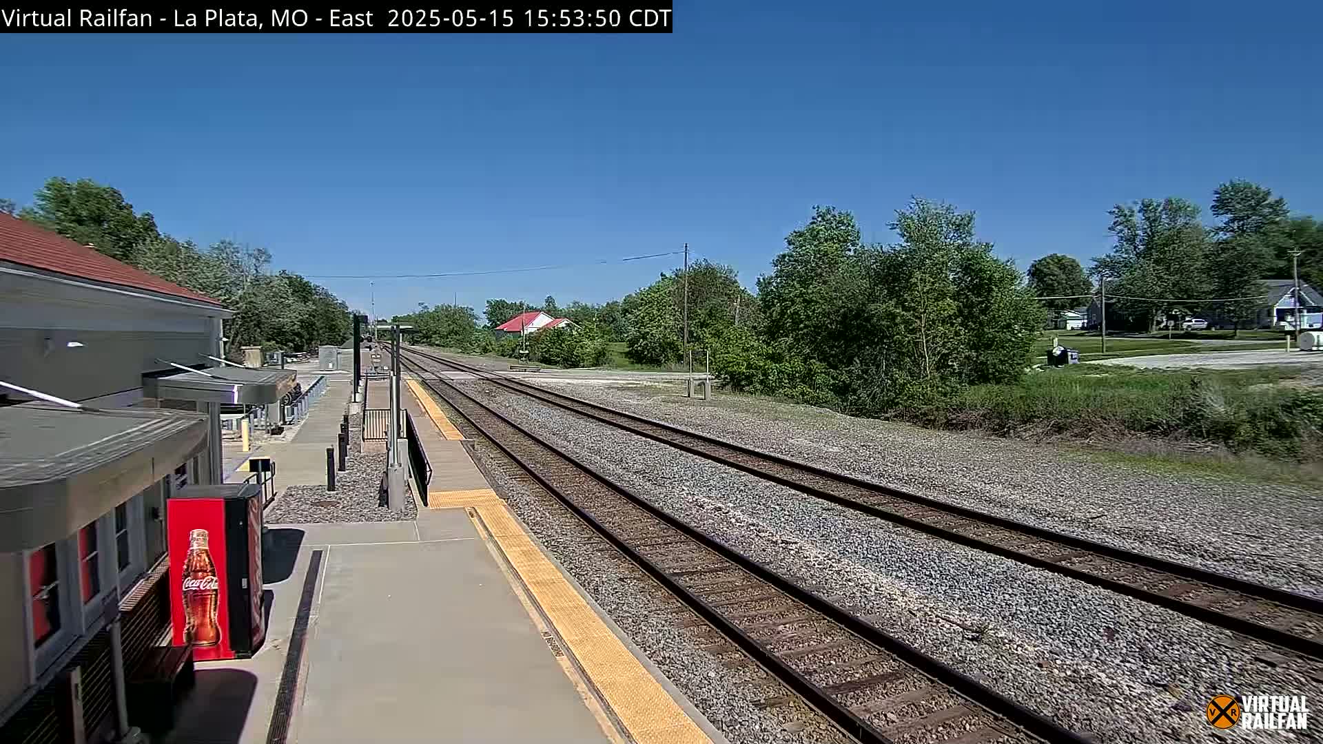 A train station platform with two parallel train tracks stretches into the distance under a clear, sunny sky, surrounded by lush green trees and vegetation.