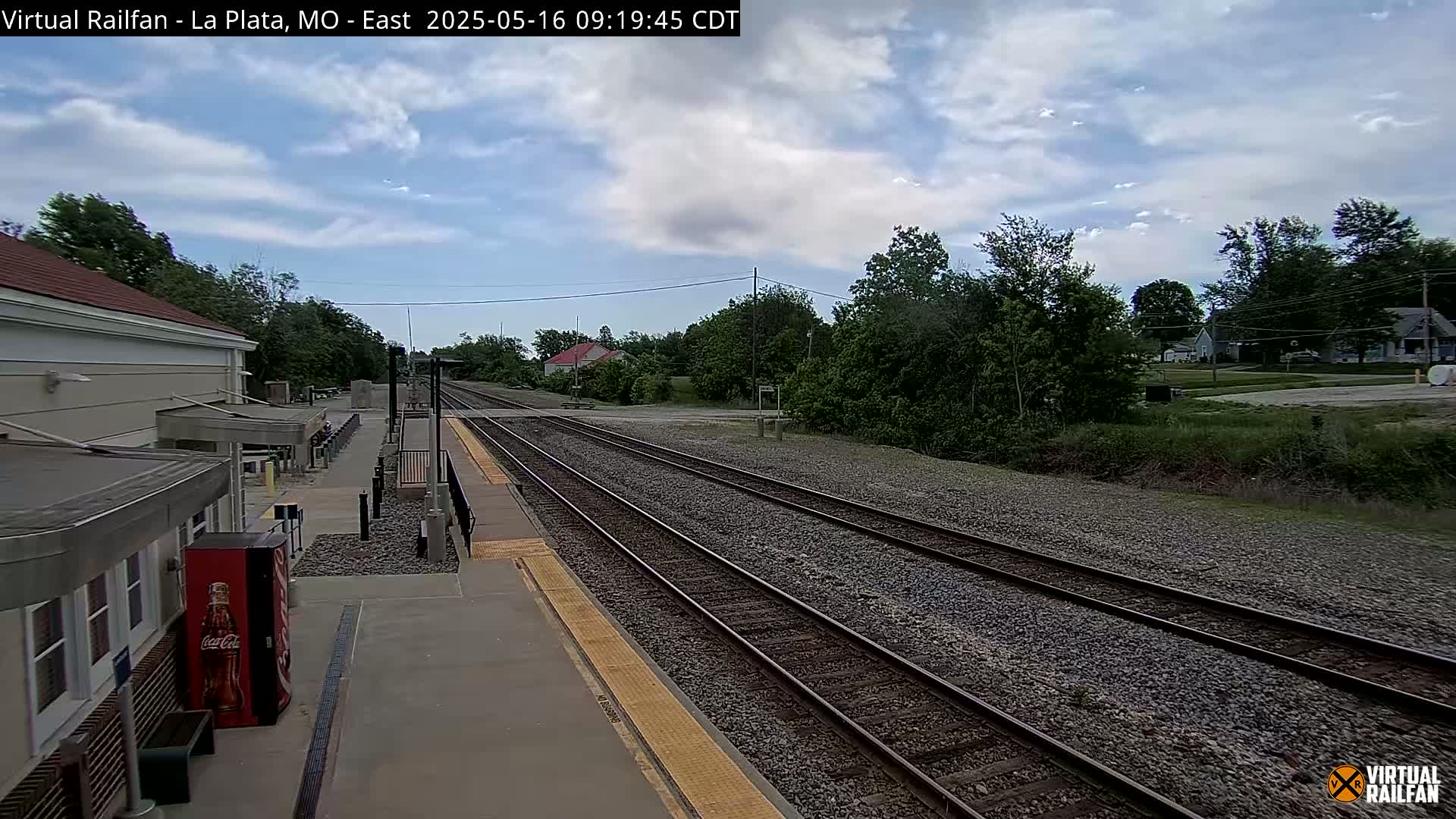A mostly sunny day at a train station shows multiple train tracks extending into a residential area with trees and houses visible in the distance.