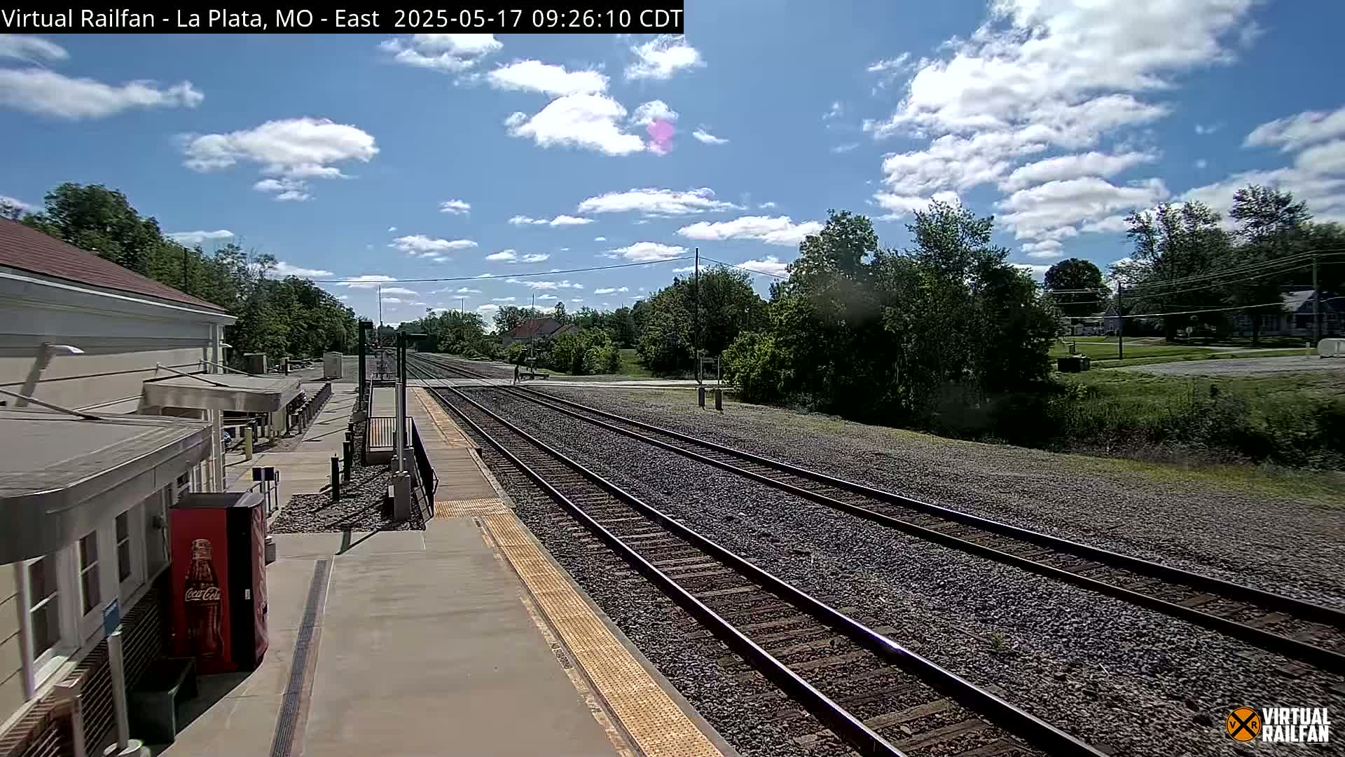 A mostly sunny day at a train station with several tracks extending into the distance, bordered by green vegetation and a few buildings.