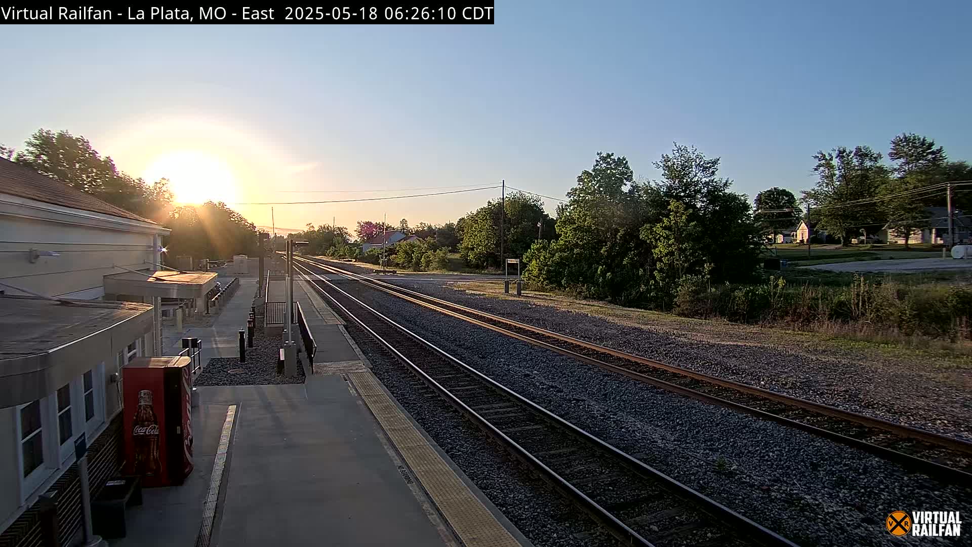 A sunrise illuminates a train station platform and two sets of railroad tracks extending into the distance, past a line of trees and houses.