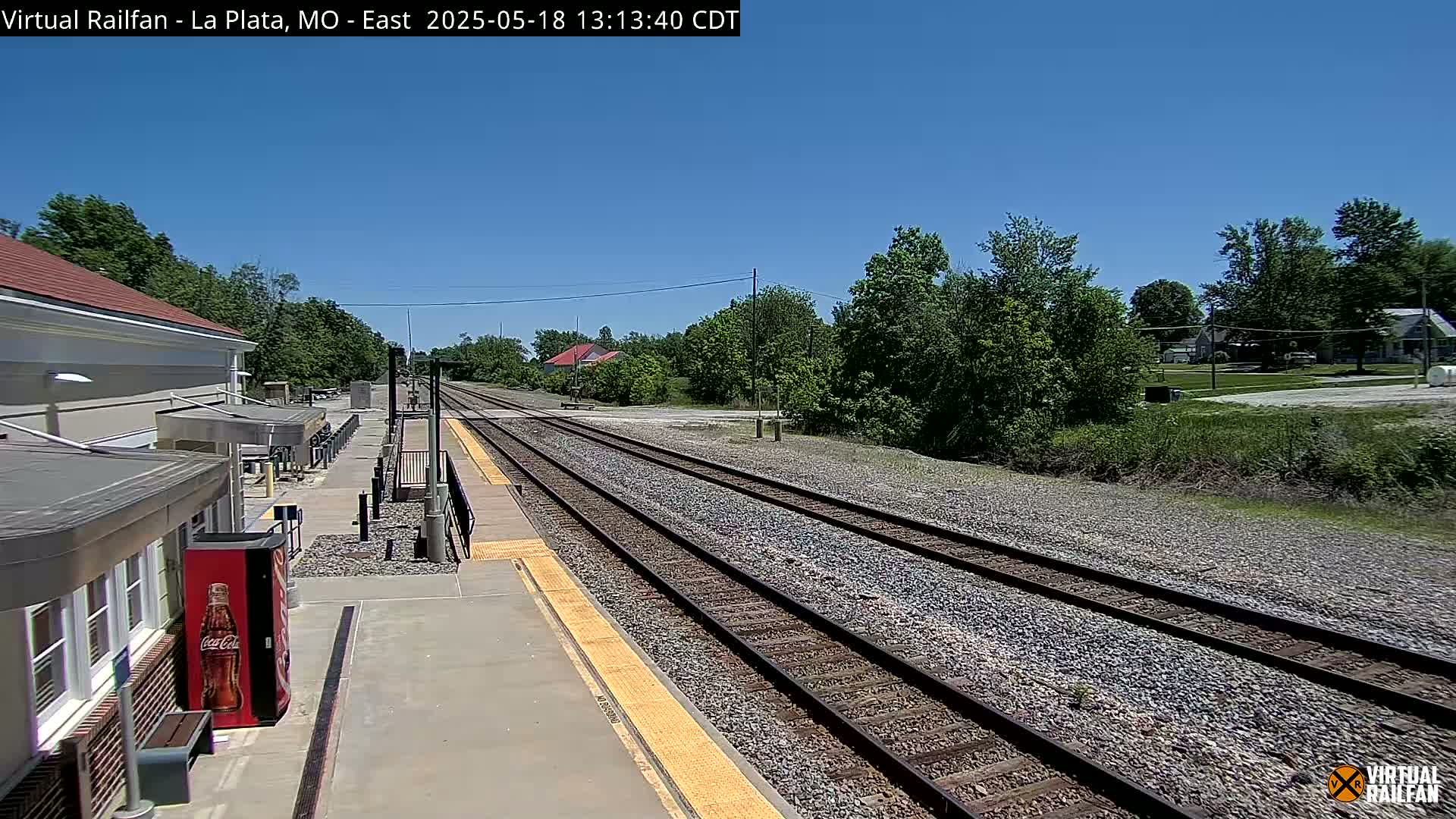 A train station platform with two train tracks extends into the distance under a clear blue sky, surrounded by green trees and vegetation.
