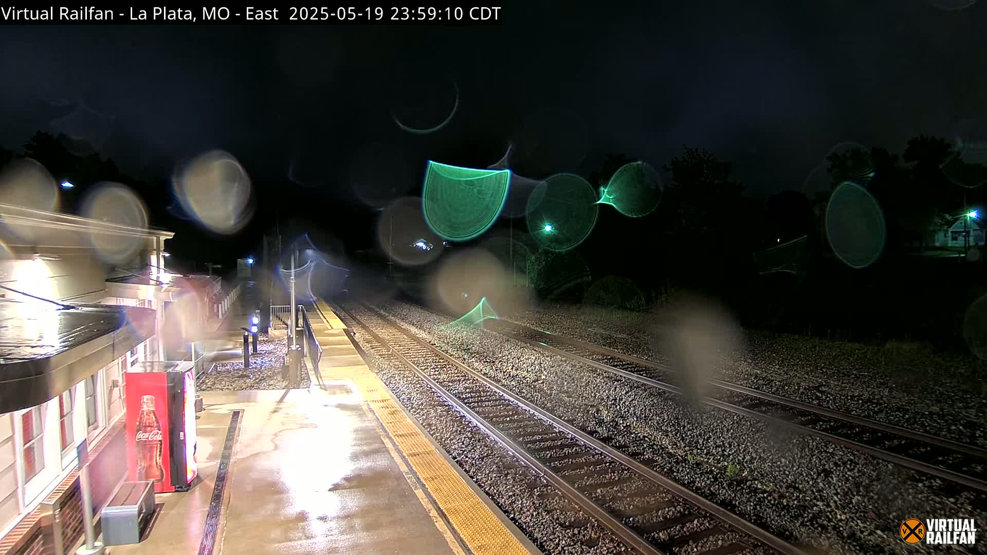 A nighttime view of a rain-slicked train platform and tracks, with raindrops obscuring the view.