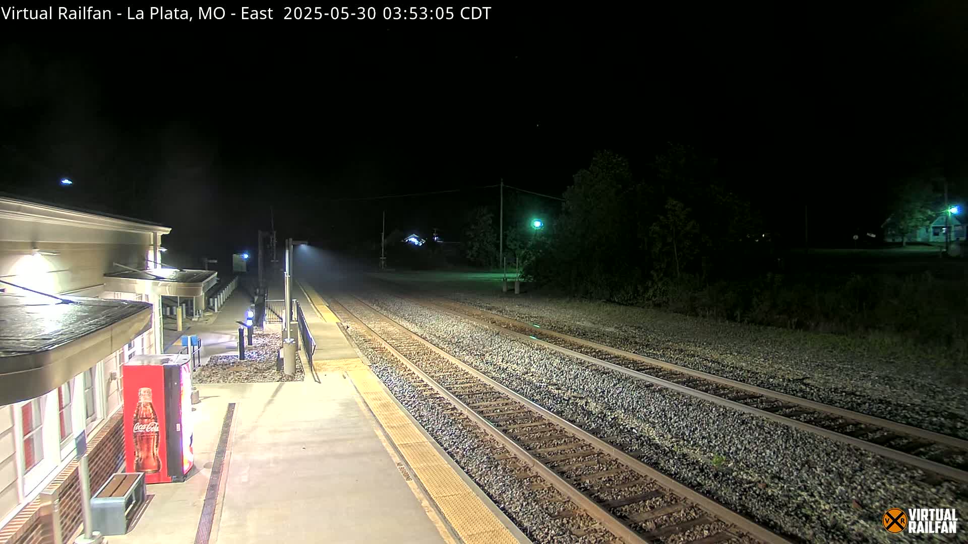 A nighttime view of a train station platform with empty train tracks extending into the darkness, illuminated by artificial lights.