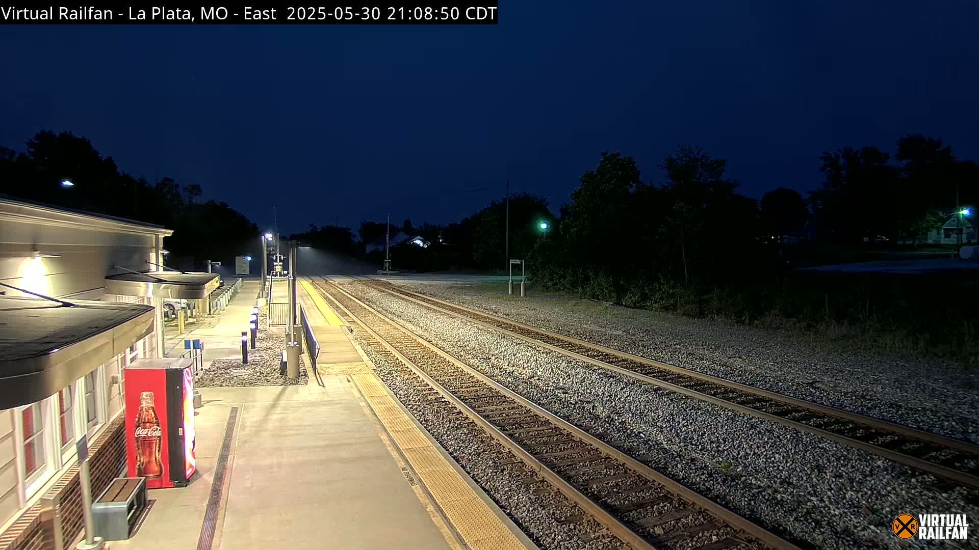 A nighttime view of a train station platform with several tracks extending into the dark distance under a clear, dark blue sky.
