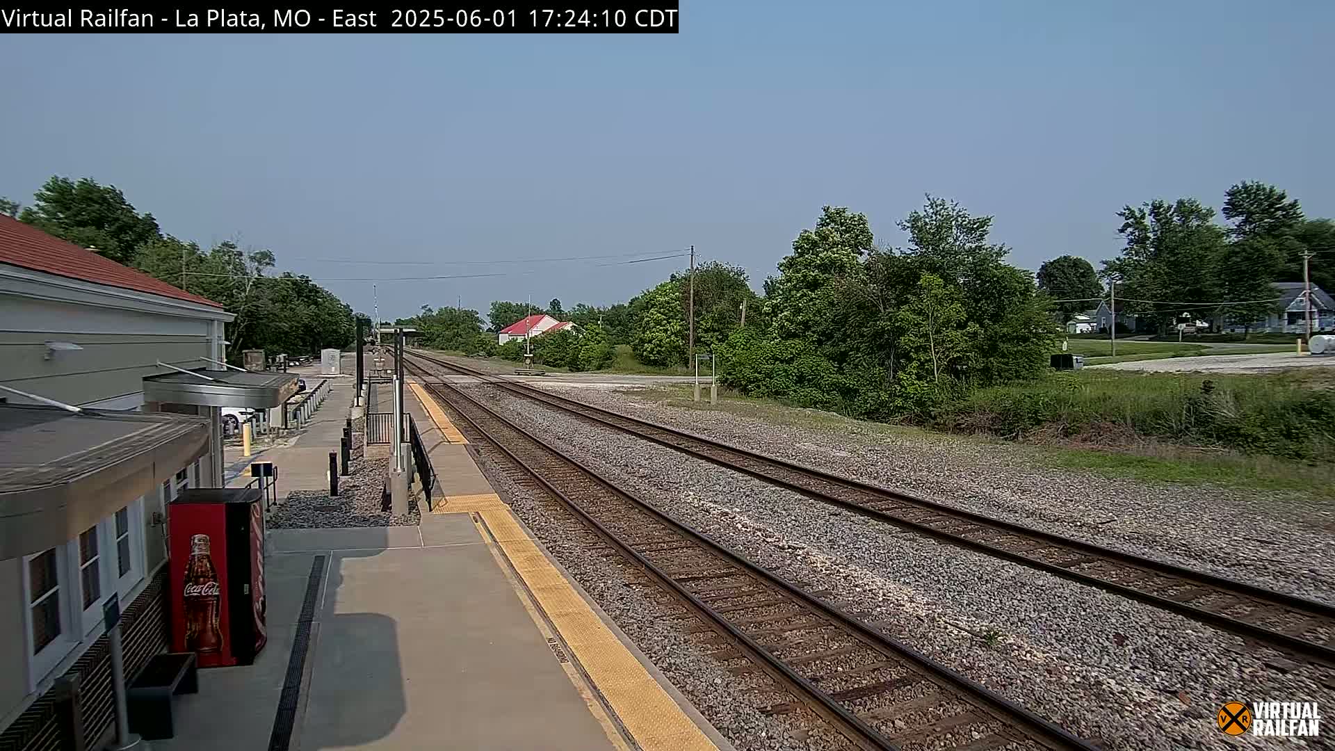 A train station platform with two sets of train tracks extends into the distance under a clear, sunny sky, surrounded by lush green trees and vegetation.