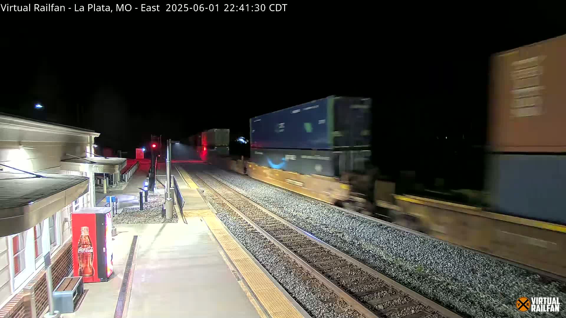 A long freight train passes a nighttime train station platform.