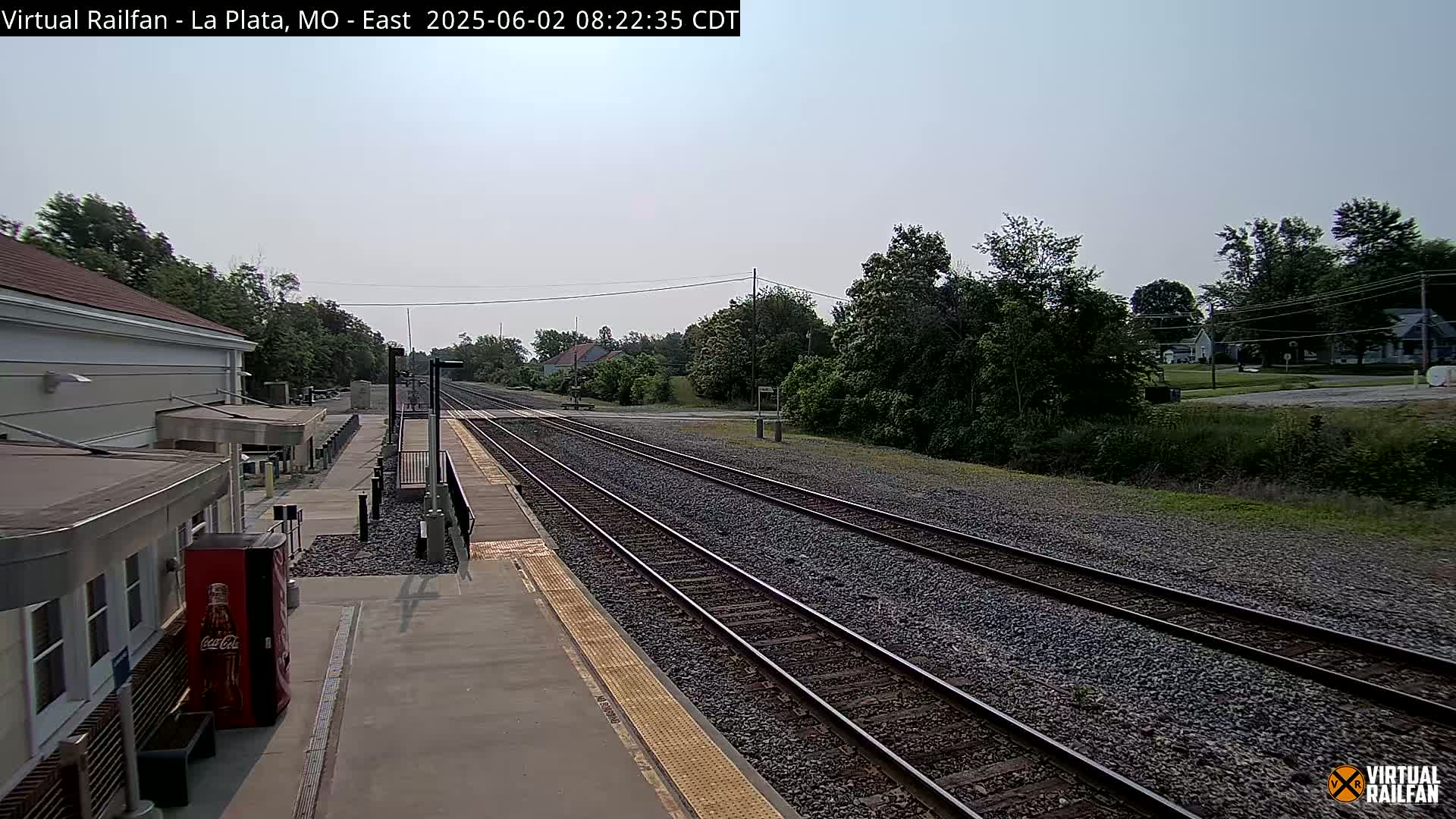 A mostly sunny view of a train station platform with multiple train tracks extending into a tree-lined landscape.
