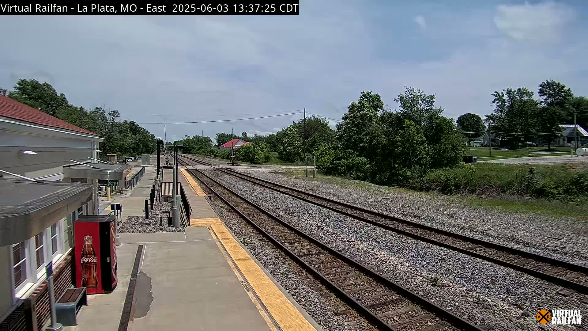 A train station platform with a view of multiple train tracks extending into a partly sunny landscape under a mostly clear sky.