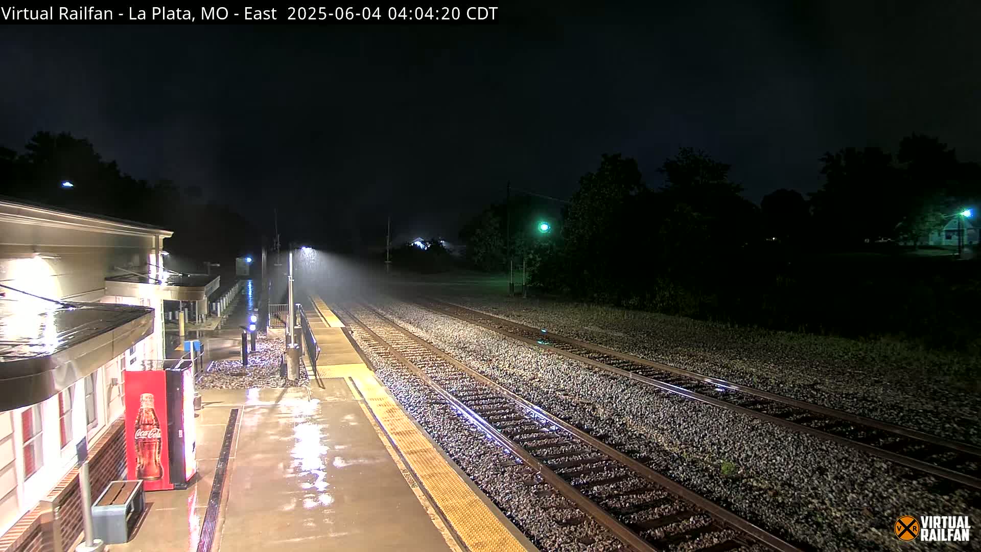 A nighttime view of a rain-slicked train platform and tracks, illuminated by artificial lights under a dark, stormy sky.