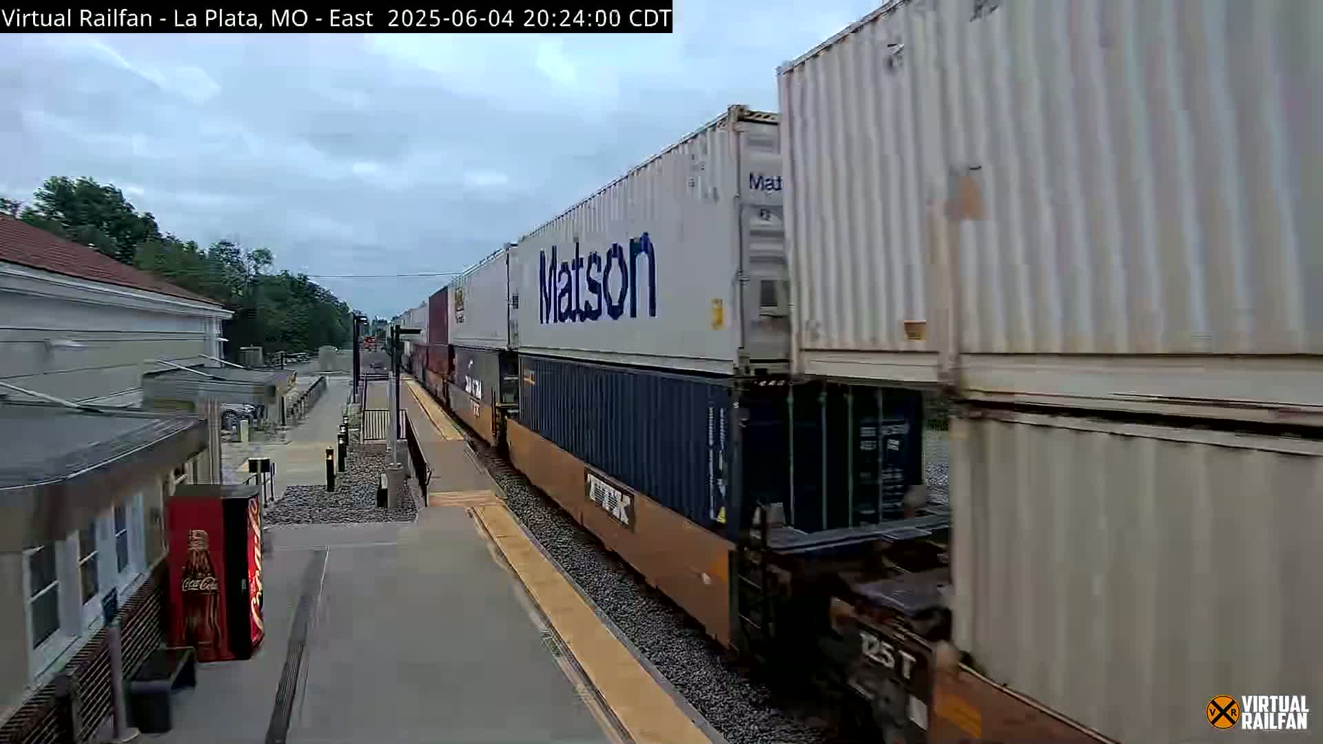 A long freight train carrying shipping containers passes a station under an overcast sky.