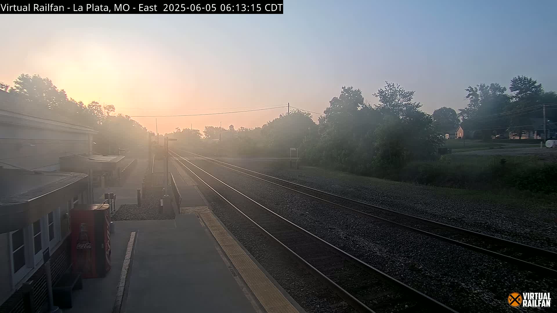 A foggy sunrise illuminates a train station platform and multiple train tracks extending into the distance.