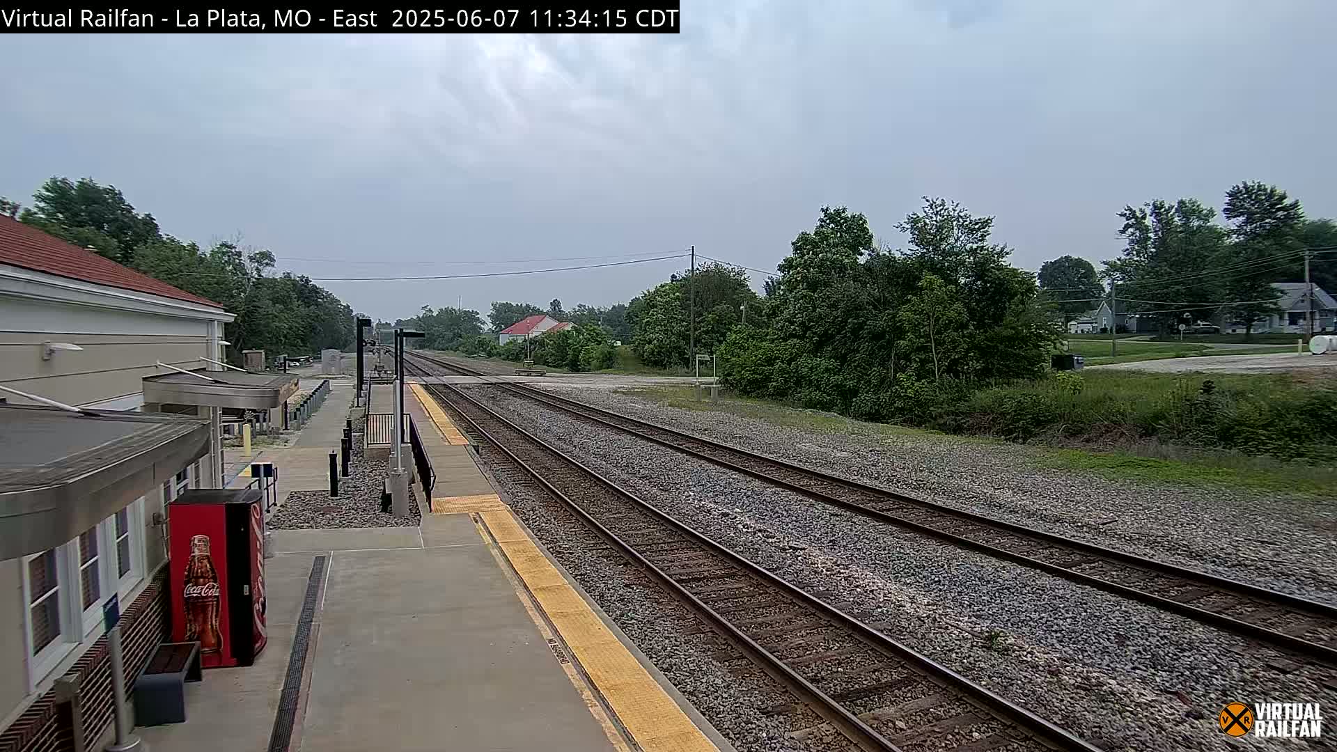 A train station platform with multiple train tracks stretches into a landscape of trees and houses under an overcast sky.