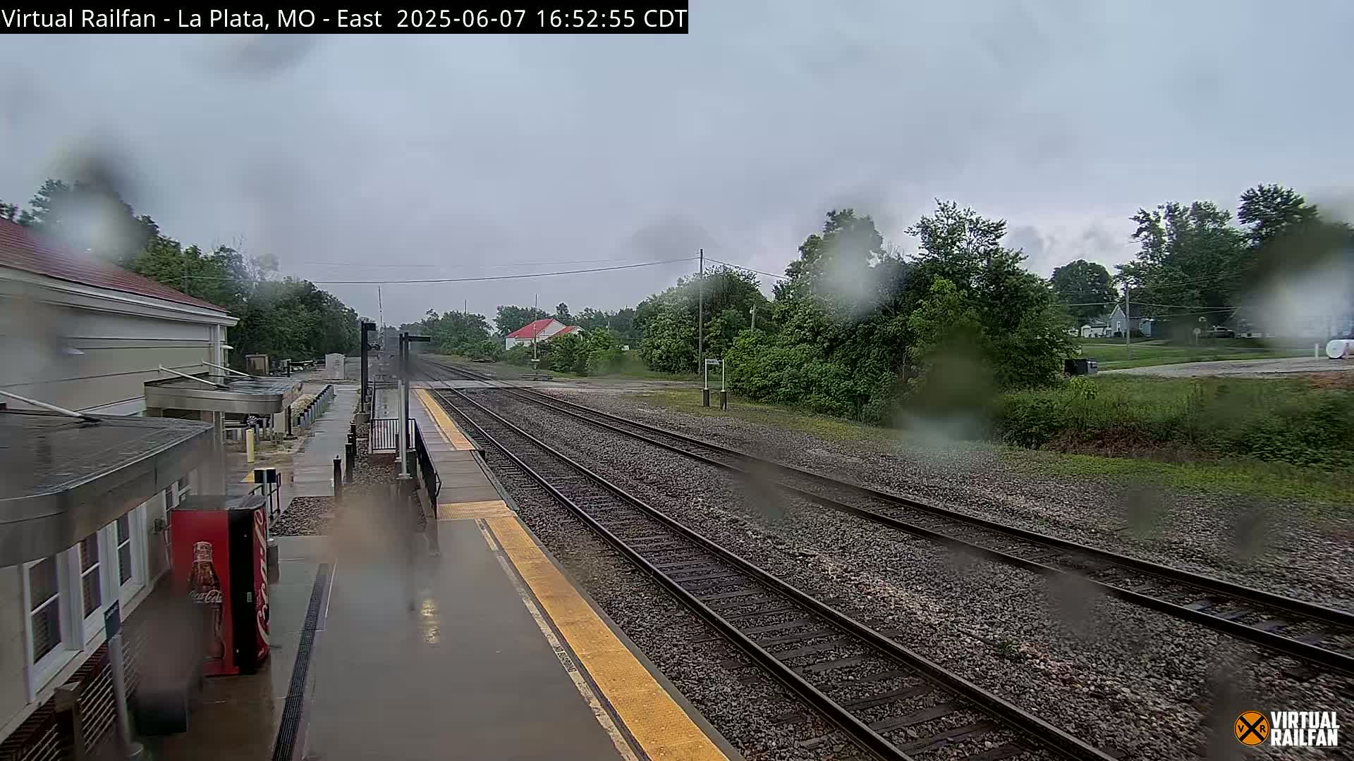 A rain-covered view of a train station platform with multiple train tracks extending into the distance under a cloudy sky.