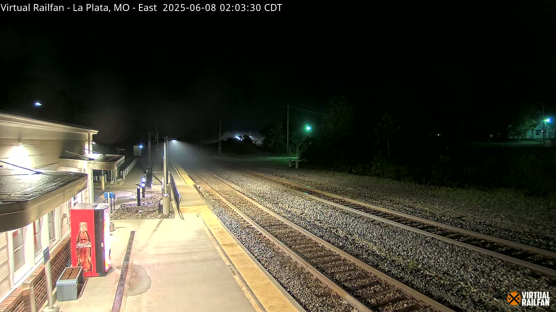 A nighttime view of a deserted train platform next to a set of train tracks under light rain.
