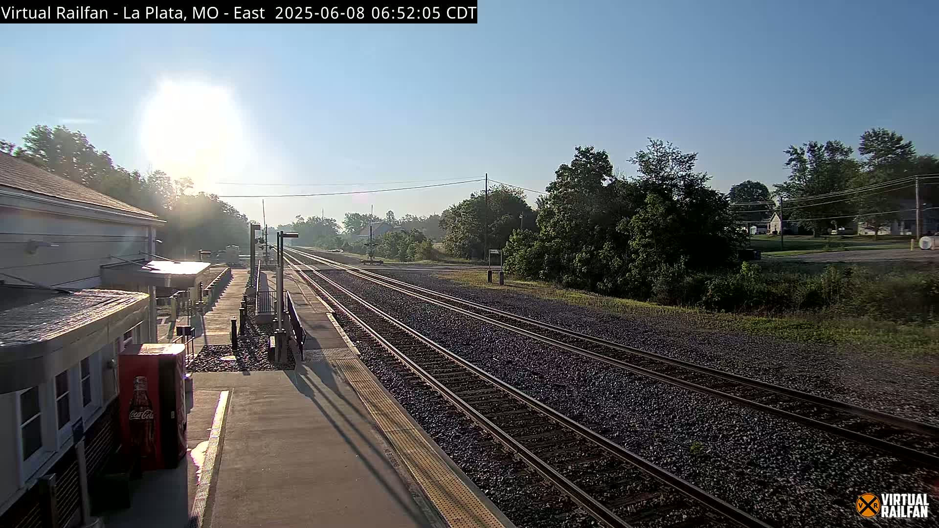 A sunny morning view of a train station platform with several train tracks extending into the distance, surrounded by green trees and vegetation.