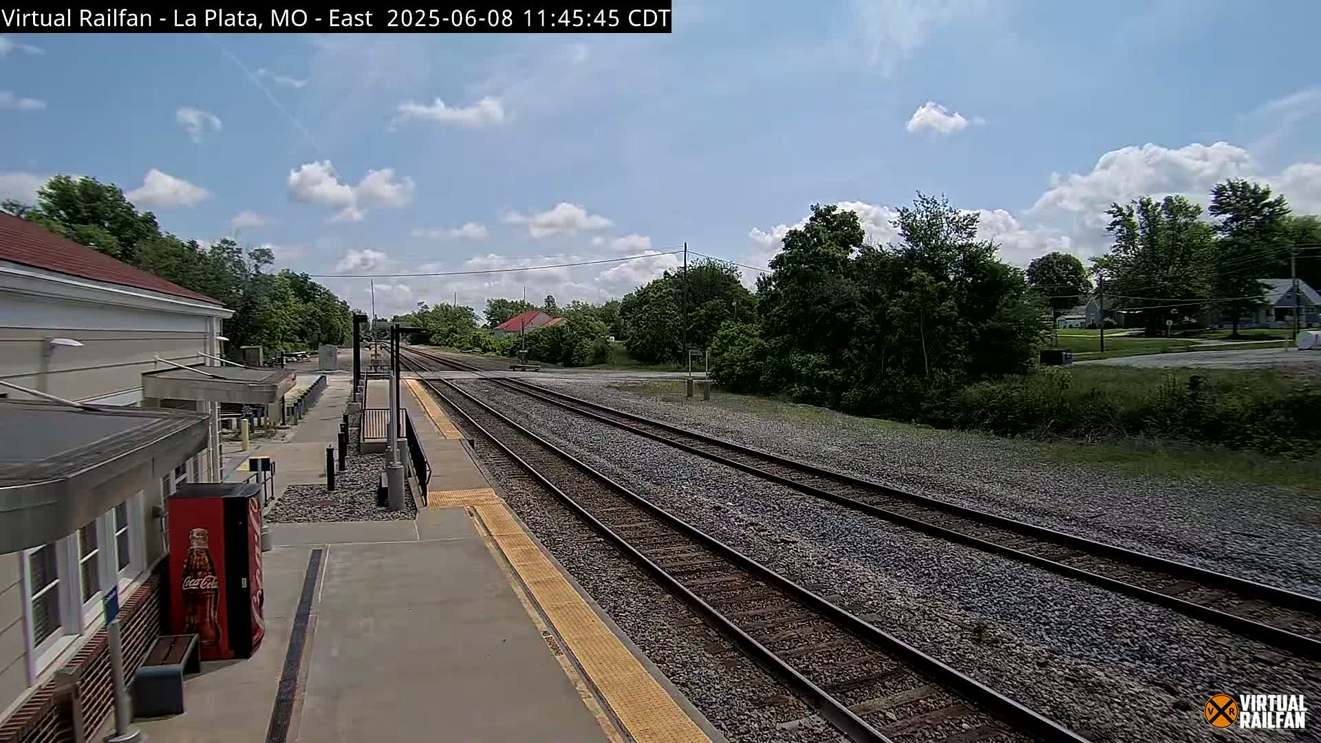 A mostly sunny day shows a train station platform with multiple train tracks extending into the distance, surrounded by lush green trees and houses.