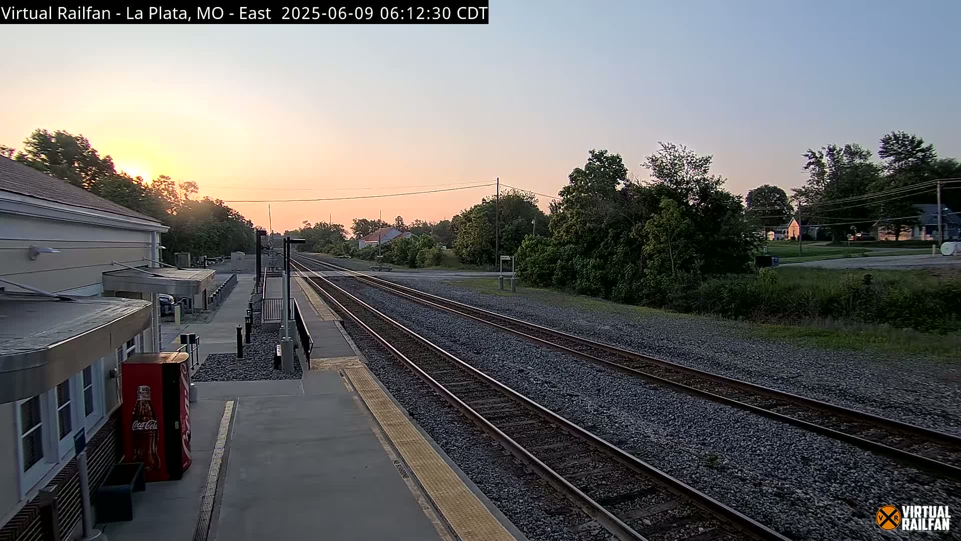 A sunrise illuminates a train station platform with multiple train tracks extending into a residential area.